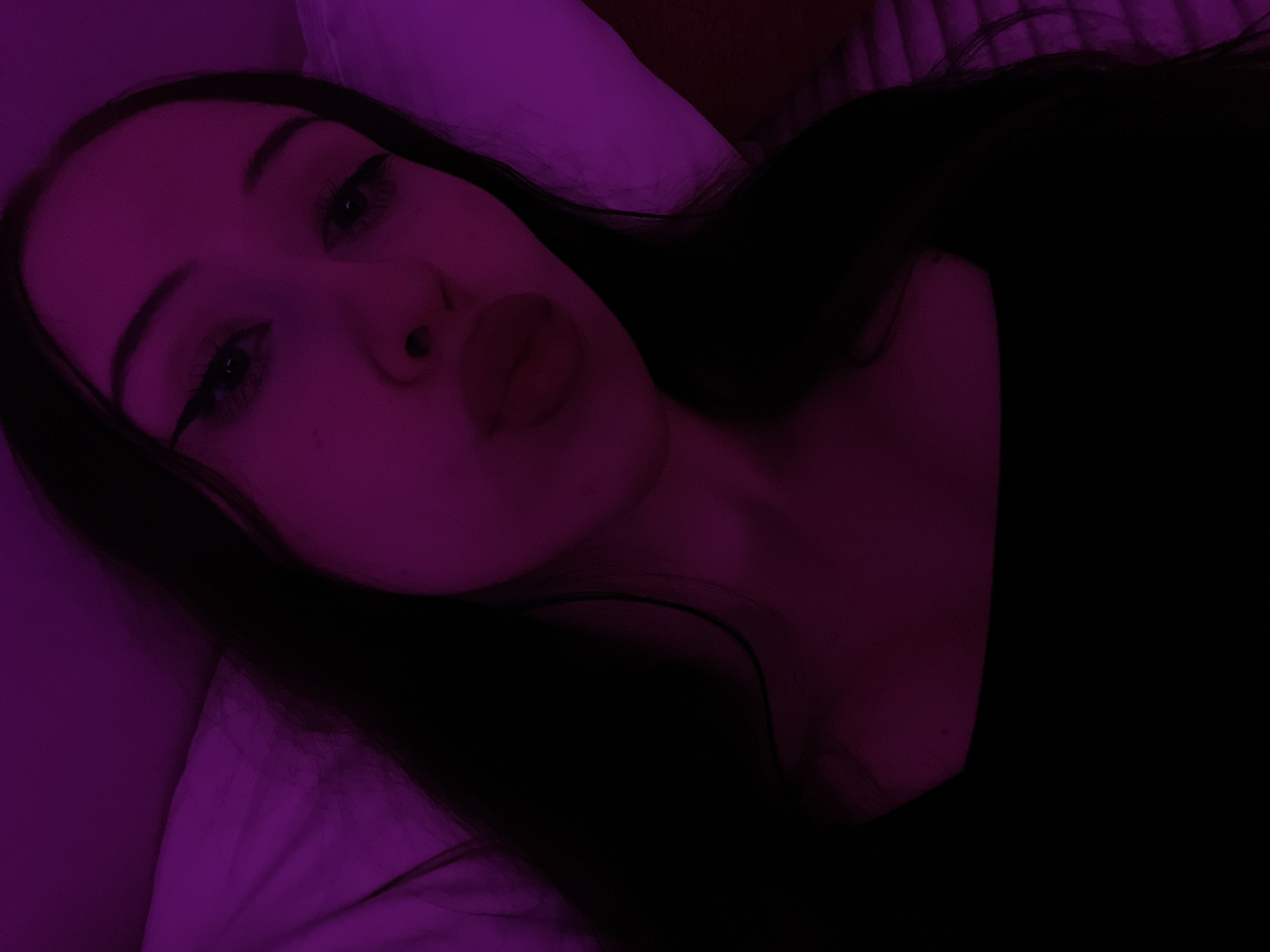 The image is a close-up selfie of a young woman with long, dark hair. She is wearing a black top. Her eyes are lined with black eyeliner and she is puckering her lips. The lighting in the image is predominantly purple, suggesting the use of colored lights or filters. The background appears to be a bed with a quilted headboard, illuminated by the same purple light. There is no identifiable information about the location in Votkinsk, Russia.