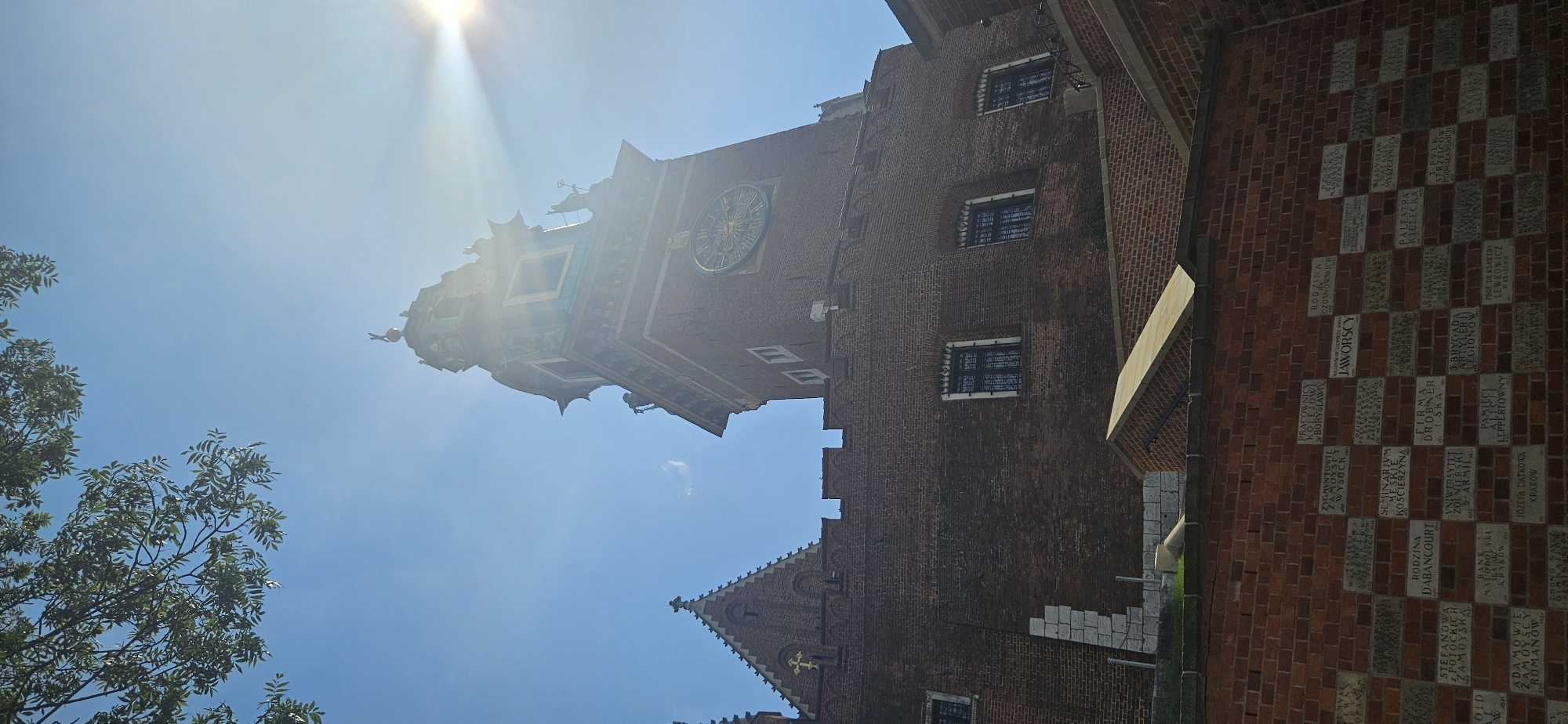 This is a vertical, low-angle shot of a brick tower and adjacent buildings in Krakow, Poland, under a bright blue sky with the sun glaring from the upper right. The tower features a large clock face with Roman numerals and intricate gold hands, and is adorned with statues and decorative architectural elements. Below the tower, a brick wall is covered with numerous stone plaques, each bearing inscriptions in Polish, likely commemorative or historical in nature. The overall setting suggests a historical or significant landmark within the city.