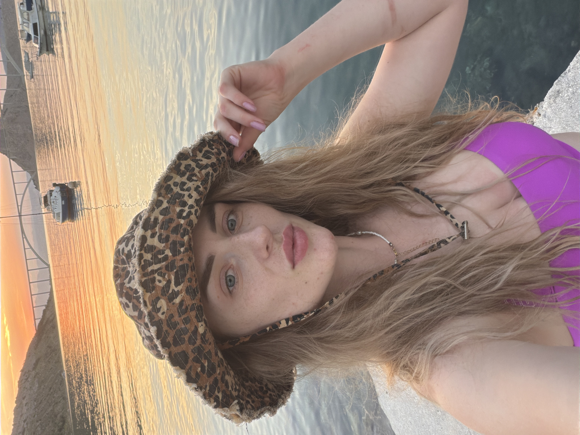 A woman is taking a selfie by the water at sunset. She is wearing a leopard-print bucket hat and a bright pink bikini top. Her blonde hair is long and wavy. She is wearing a necklace with beads and a pendant. Her left hand is resting on the brim of her hat, and her right arm is bent at the elbow, with her hand near her head. There are some red marks on her right arm.
The background shows a body of water with several boats. In the distance, a bridge spans across a body of water, with hills on either side. The sky is filled with the warm colors of sunset, casting a golden glow on the water and the landscape. The woman is standing on a concrete surface, possibly a pier or a dock, with the water extending into the background. The image appears to have been taken outdoors, in a coastal or waterfront location.