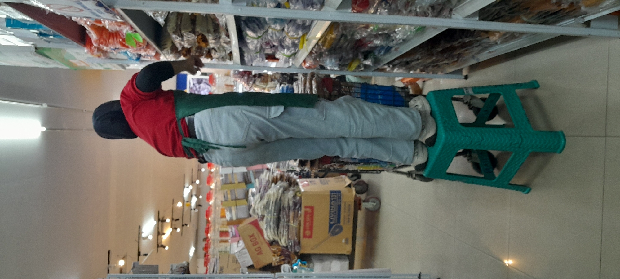 In Rogojampi, Indonesia, a person wearing a red shirt, a green apron, and light-colored pants stands on a teal plastic stool. The person is facing away from the camera, looking towards shelves stocked with goods. In the background, there are aisles of merchandise and lighting fixtures on the ceiling. To the left, a cart with boxes is visible. The scene appears to be inside a store or warehouse.