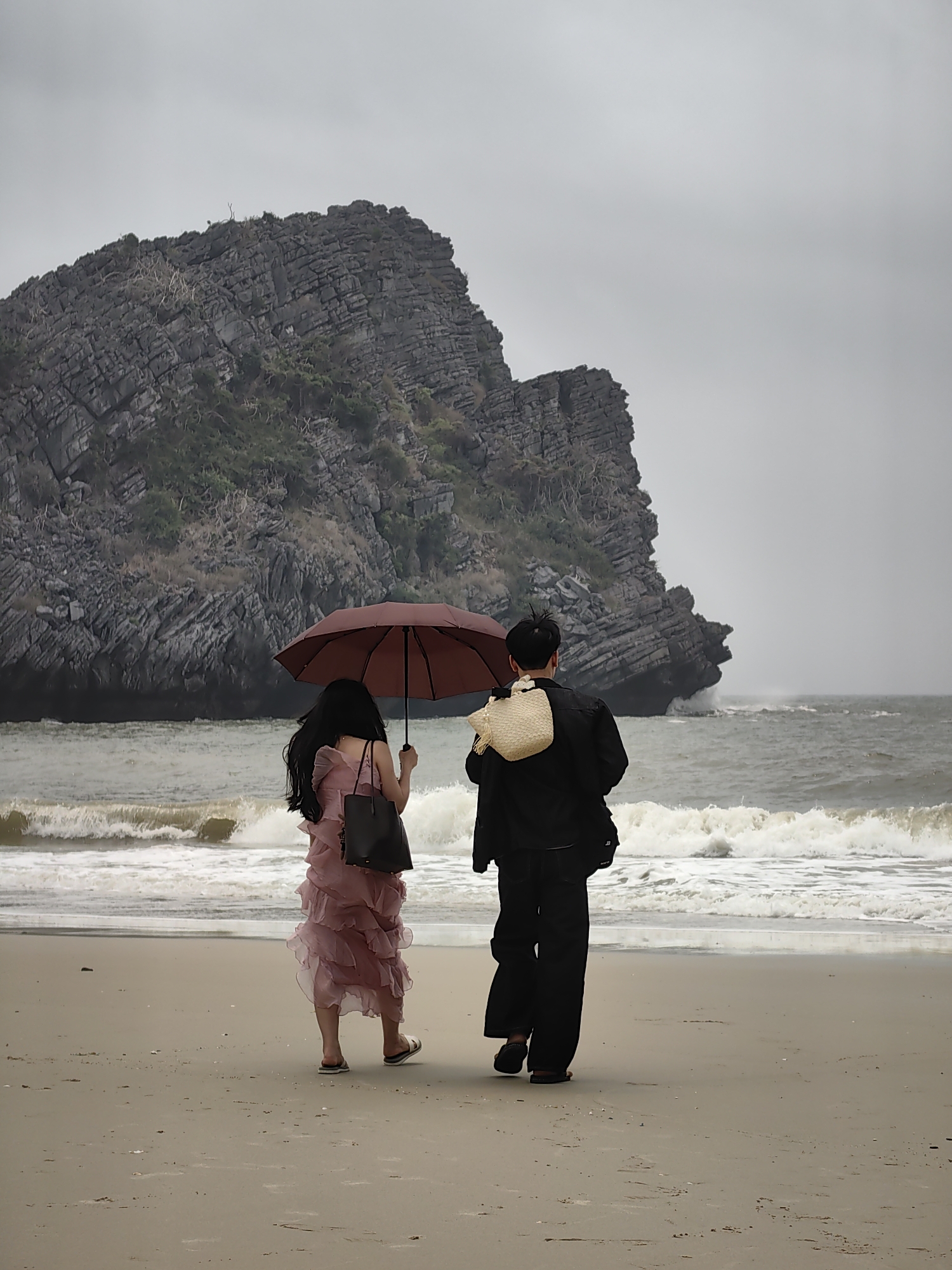 On a beach in Hải Phòng, Vietnam, a woman in a pink dress and a man in black pants and a jacket walk towards the sea. They are sharing a maroon umbrella. The man is carrying a woven straw bag on his back. A large rocky formation sits in the background of the scene. Waves are breaking on the shore. The sky is overcast.