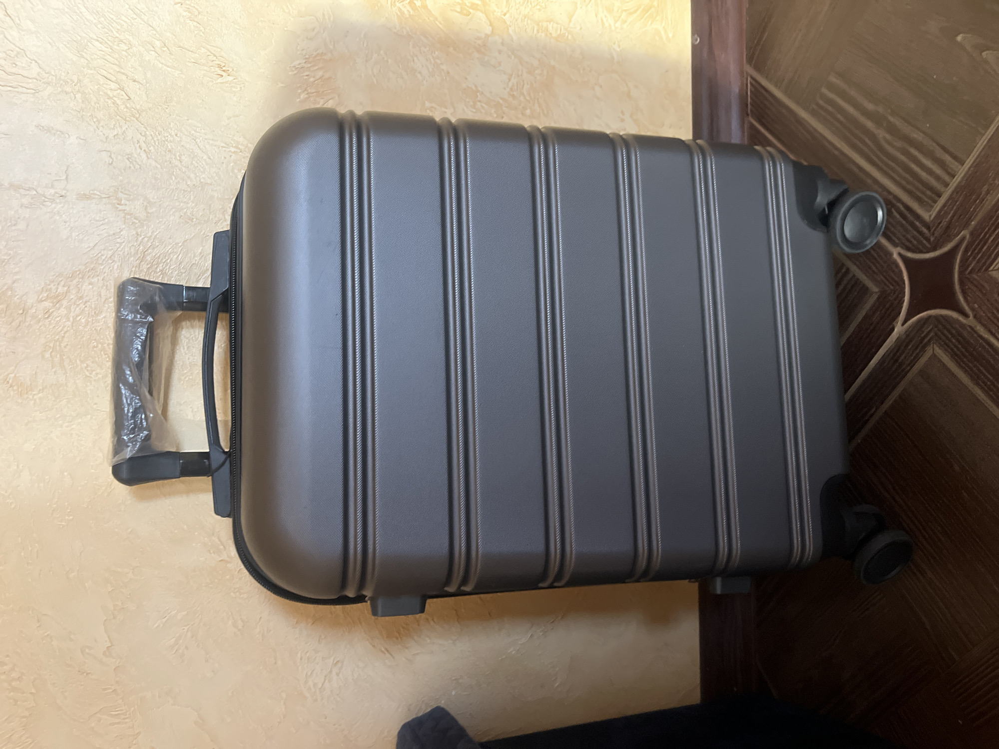 A dark grey suitcase with a plastic wrap on its handle is standing upright on a wooden floor. The suitcase has a textured surface with horizontal lines and appears to be a carry-on size with four wheels. It is positioned against a textured beige wall. To the left of the suitcase, a dark object, possibly a couch or a chair, is partially visible. The image shows the suitcase from a front, slightly angled perspective. The scene is indoors, with a wooden baseboard visible at the bottom of the wall. There is no indication of the city of Tarnivtsi, Ukraine in the image.
