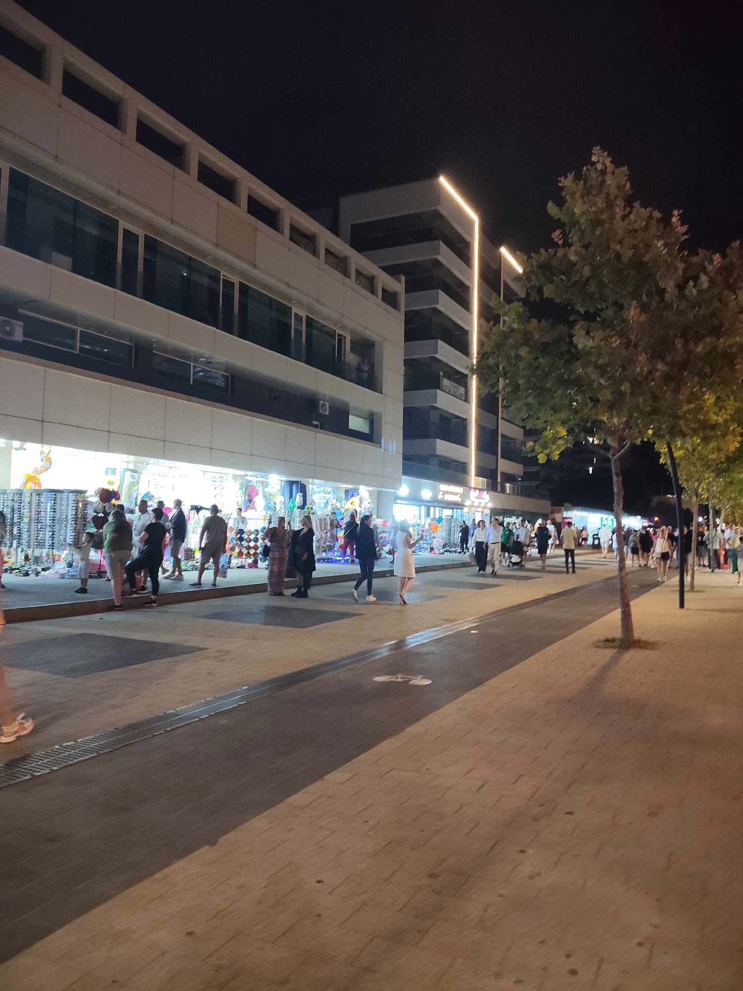 The image captures a nighttime scene on a pedestrian walkway in the city of Constanța, Romania. The walkway is lined with shops on the left and trees on the right. In the background, there are multi-story buildings, one of which features prominent modern lighting that illuminates the facade with vertical golden bands. Several people are visible on the walkway, some browsing in front of the shops, others walking or standing. The shops appear to be selling various merchandise, as suggested by displays of items like sunglasses and clothing. The ground of the walkway is paved with interlocking bricks, and streetlights cast shadows. The overall atmosphere suggests a lively commercial or tourist area.