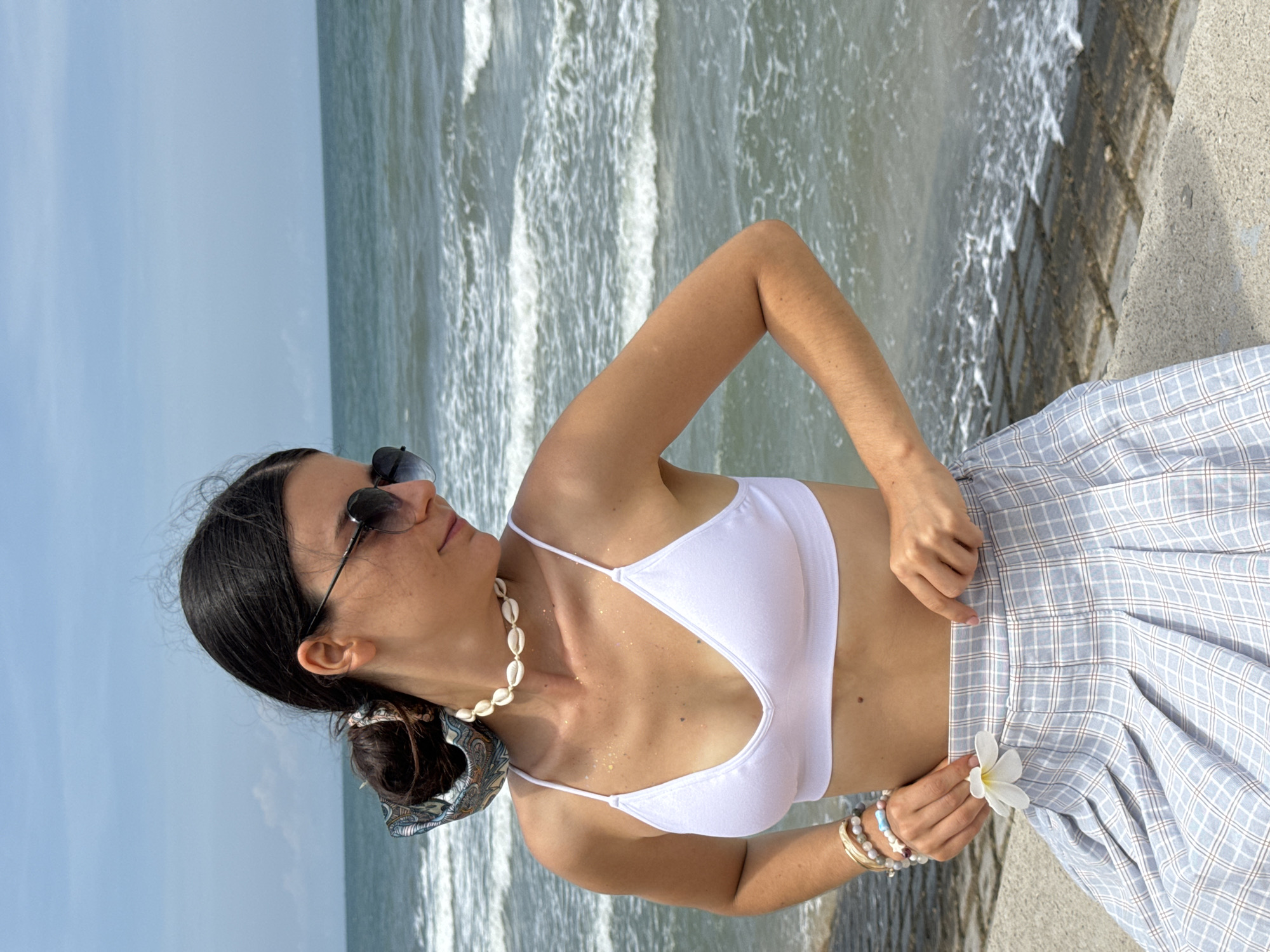 A woman with dark hair pulled back in a messy bun and wearing a patterned scarf stands at the edge of the ocean. She has sunglasses on and a shell necklace. She is wearing a white bikini top and a light blue plaid skirt. Her right arm is bent, with her hand resting on her hip, and her left hand is pulling at the waistband of her skirt, where a white flower is tucked. The background shows a cloudy sky and the ocean with small waves breaking on the shore. The woman is standing on a concrete embankment, with patterned concrete slabs visible behind her. This scene is located in Phu Thuy Ward, Vietnam.