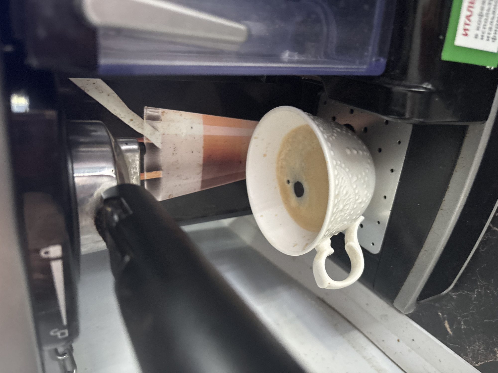 This is a close-up, eye-level, indoor photograph of an espresso machine with a freshly brewed cup of coffee. The espresso machine is dark gray and black, with a silver portafilter visible at the top. A clear plastic container, likely for water or milk, is situated to the right of the machine. A white ceramic cup, decorated with a subtle embossed pattern, sits on a black drip tray beneath the portafilter, filled with light brown coffee topped with a darker crema. A translucent plastic cup, containing a reddish-brown liquid with froth, is positioned behind the white coffee cup and to the left of the clear container. The overall environment appears to be a kitchen or countertop, with a light-colored surface below the machine and a dark, patterned surface beneath the drip tray. There are no people visible in the image. The photograph does not provide any specific information about its location in Sochi, Russia.