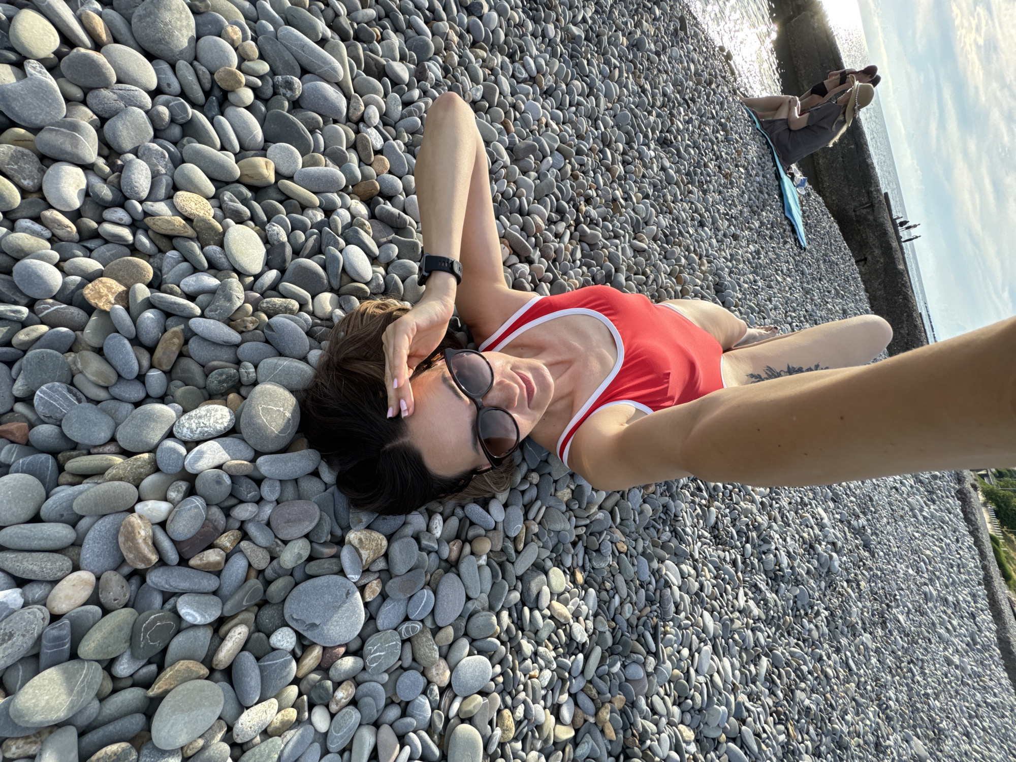 A woman in a red swimsuit lies on a pebble beach in Sochi, Russia, taking a selfie. She wears sunglasses and a black watch. In the background, several people sit on a concrete embankment near the water, and a blue towel is visible on the pebbles. The sky is partly cloudy.