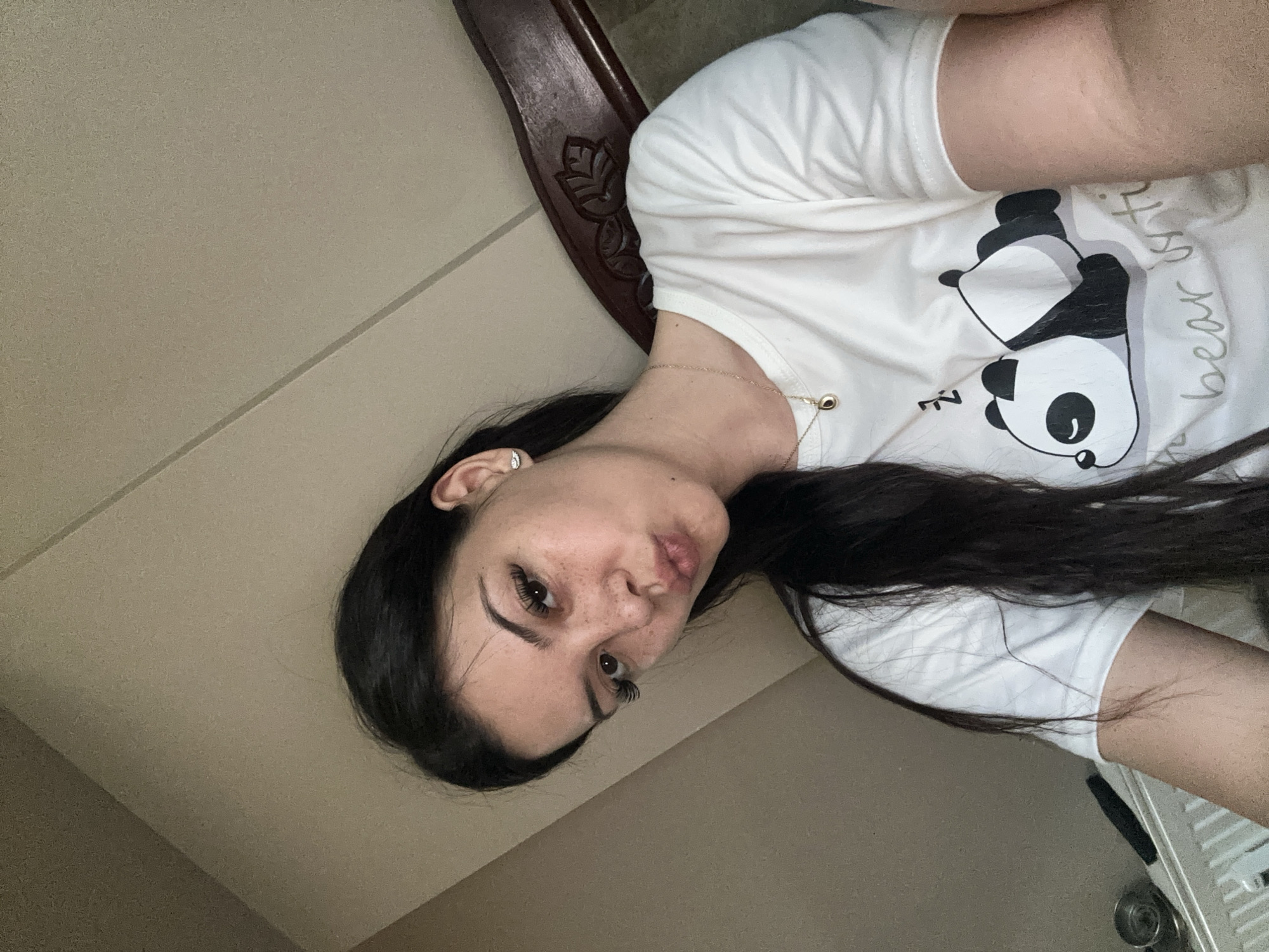 A young woman with long, dark hair and freckles is taking a selfie. She is wearing a white t-shirt with a cartoon panda design. The background appears to be a room with beige walls and a wooden headboard. There are no visible landmarks or indications of the city of Sliven, Bulgaria.