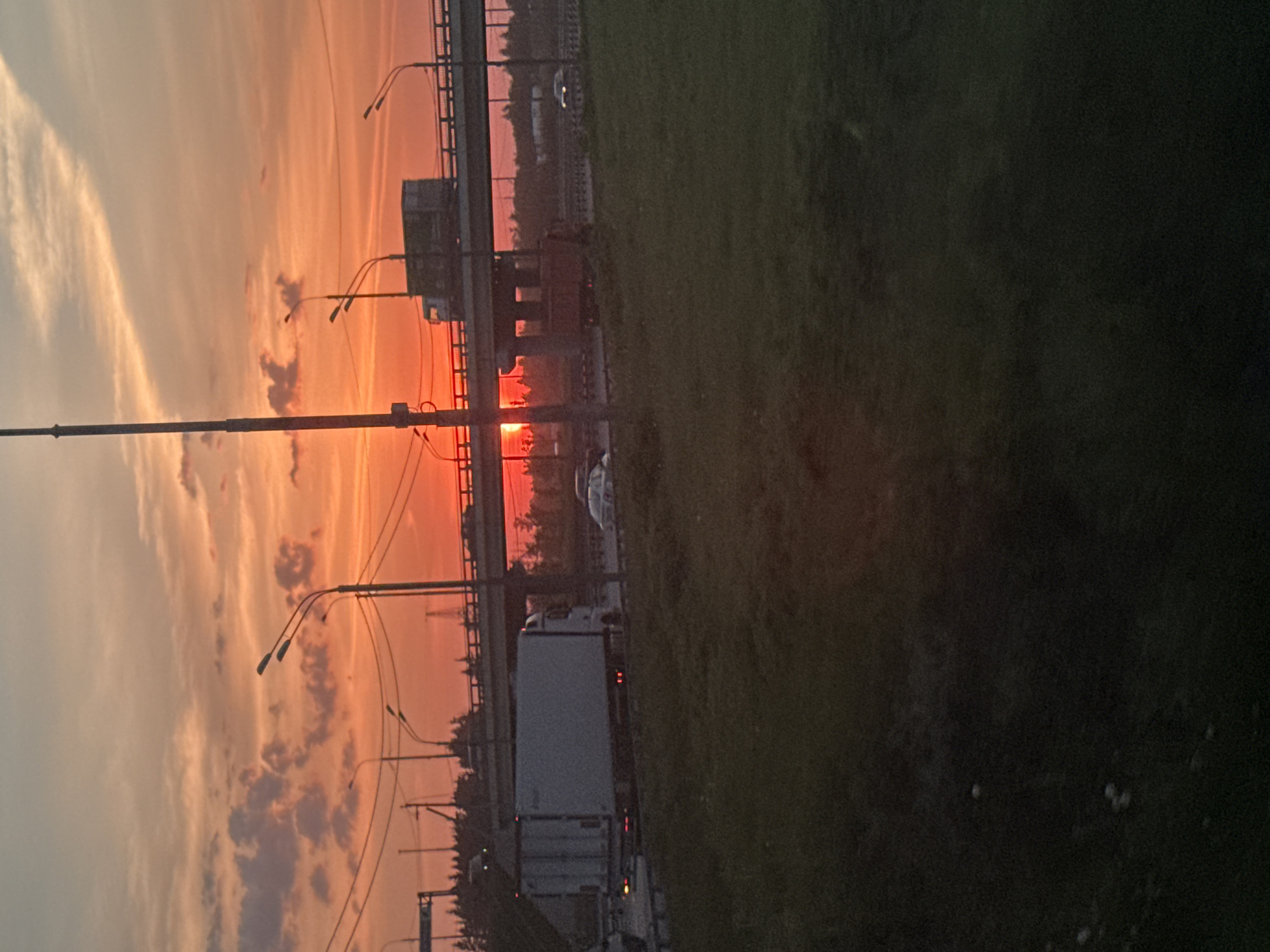 The image shows a sunset in посёлок опытного хозяйства «Ермолино», Russia. A highway overpass with streetlights is visible. There is a truck on the road below and a car on the highway above. The sky is filled with orange and yellow clouds. The grass is green.