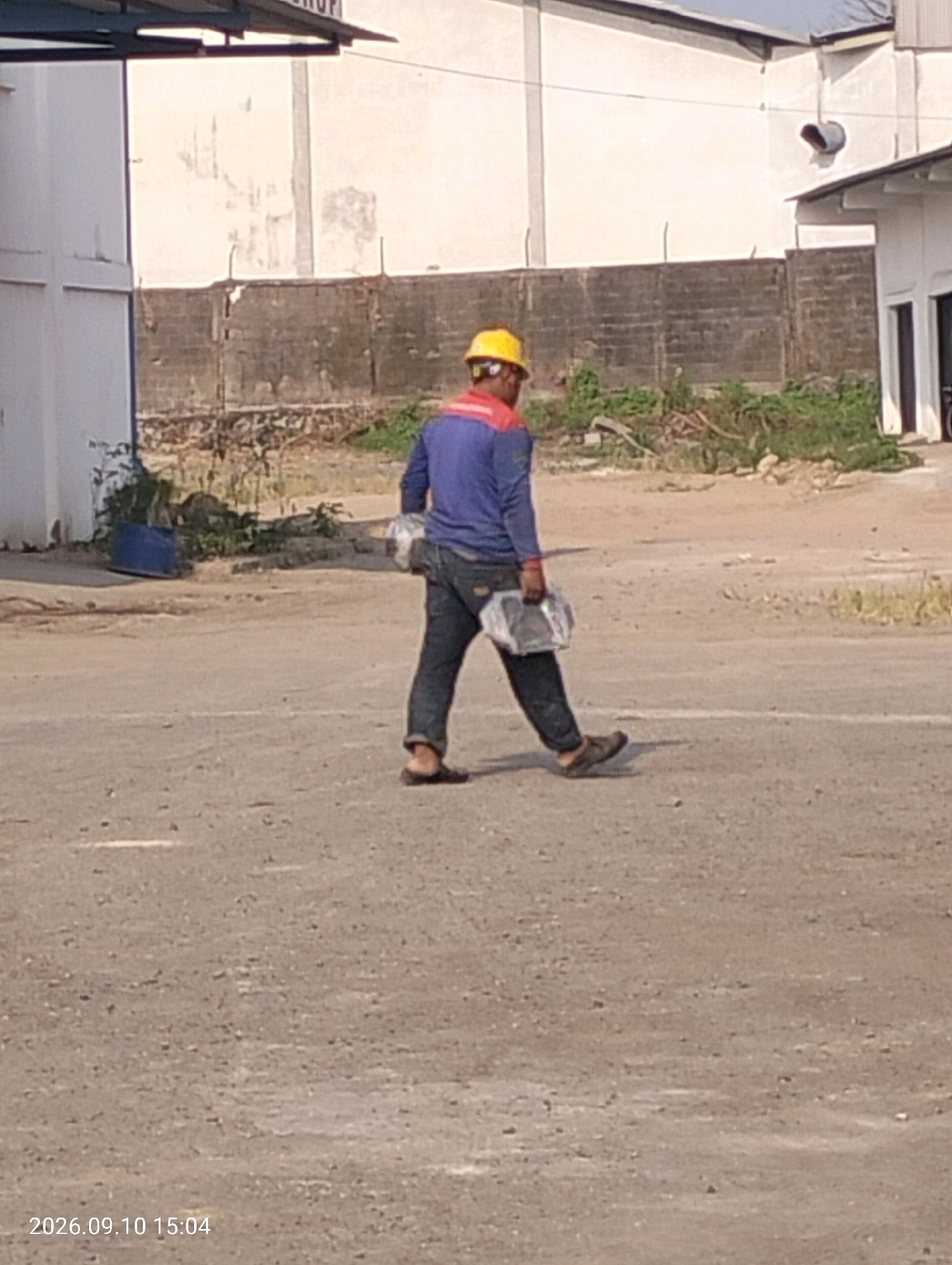 A person wearing a yellow hard hat and a blue long-sleeved shirt with red accents on the collar and shoulders walks across a dirt ground. The person is also wearing dark pants and dark open-toed shoes. In their right hand, they are carrying a clear plastic bag containing what appears to be food. The person is walking away from the camera towards the right side of the frame. In the background, there is a white building with a dark-colored roof and a section of a brick wall. The environment appears to be an industrial or construction area. The timestamp on the image indicates it was taken on September 10, 2026, at 15:04. The location is identified as Bambe, Indonesia.
