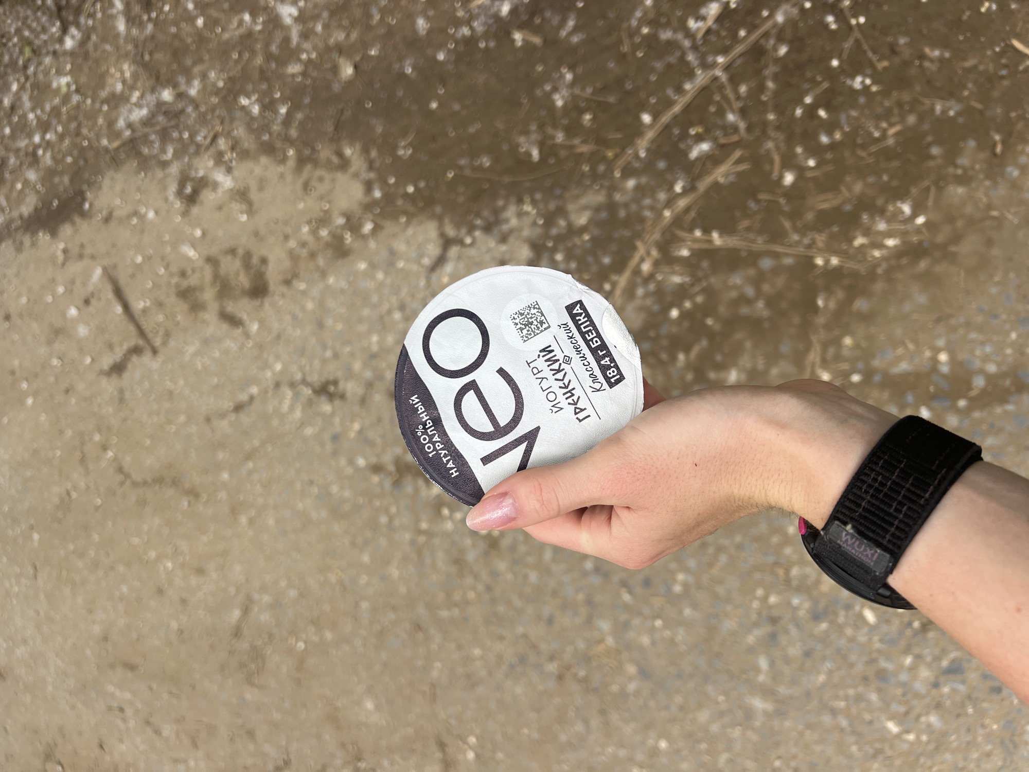 A human's right hand holds a round container of NEO Greek yogurt, centered in the frame. The yogurt lid, white with black text, displays "100% НАТУРАЛЬНЫЙ" (100% Natural), the brand "NEO," "ЙОГУРТ ГРЕЧЕСКИЙ Классический" (Greek Yogurt Classic), "18,4 г БЕЛКА" (18.4g protein), and a QR code. The individual's fingernails are shaped and covered with a light-colored, possibly shimmery, polish. A black fabric wristband, identified by a small "WUXI" tag and a visible pink button on the watch body, is worn on the wrist. The background is an out-of-focus outdoor ground, consistent with a dirt or gravel path, exhibiting shades of light brown and tan, scattered small debris, and some dry plant matter. The setting is in the city of Chaikovsky, Russia.