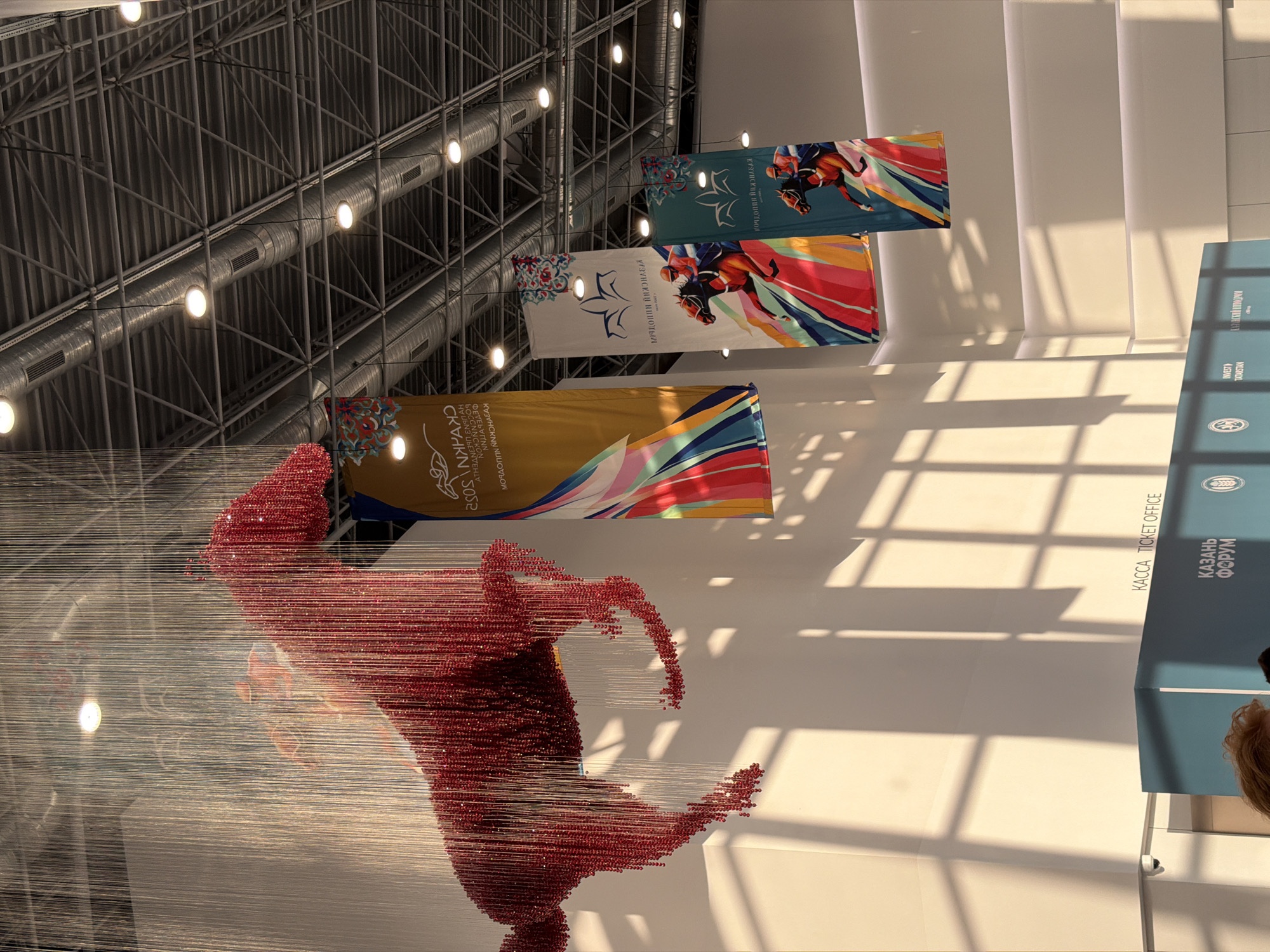 A low-angle, indoor shot captures a red beaded sculpture of a horse suspended from the ceiling. The sculpture is made of numerous small, red beads threaded onto thin wires, creating a shimmering, almost ethereal effect. The horse is depicted in a galloping pose, its body formed by the dense beading and its mane and tail trailing downwards. Directly behind the sculpture, a white wall casts a shadow in the shape of a window pane.
Hanging vertically from the ceiling are three large banners. The leftmost banner is gold and features a colorful, abstract design with streaks of pink, orange, yellow, blue, and green. The middle banner is white with a dark blue border and depicts two jockeys on horseback in mid-race, rendered in a bold, graphic style against a background of colorful abstract shapes. The rightmost banner is teal with a dark blue border and also shows two jockeys on horseback, similar in style to the middle banner. Text is visible on all three banners, in a language that appears to be Cyrillic.
Above the banners, the industrial ceiling is visible, with exposed metal beams, air ducts, and circular light fixtures. Sunlight streams in from an unseen source, creating strong diagonal shadows on the white wall.
In the lower portion of the image, a teal-colored structure with "КАЗАНЬ ФОРУМ" printed in white lettering is partially visible. Next to this, the text "KACCA TICKET OFFICE" is written in black on the white wall. A small security camera is mounted on the left side of the wall. The hair of a person with auburn hair is visible in the bottom left corner, suggesting their presence within the scene. The overall environment appears to be a large, modern indoor space, possibly an exhibition hall or convention center.