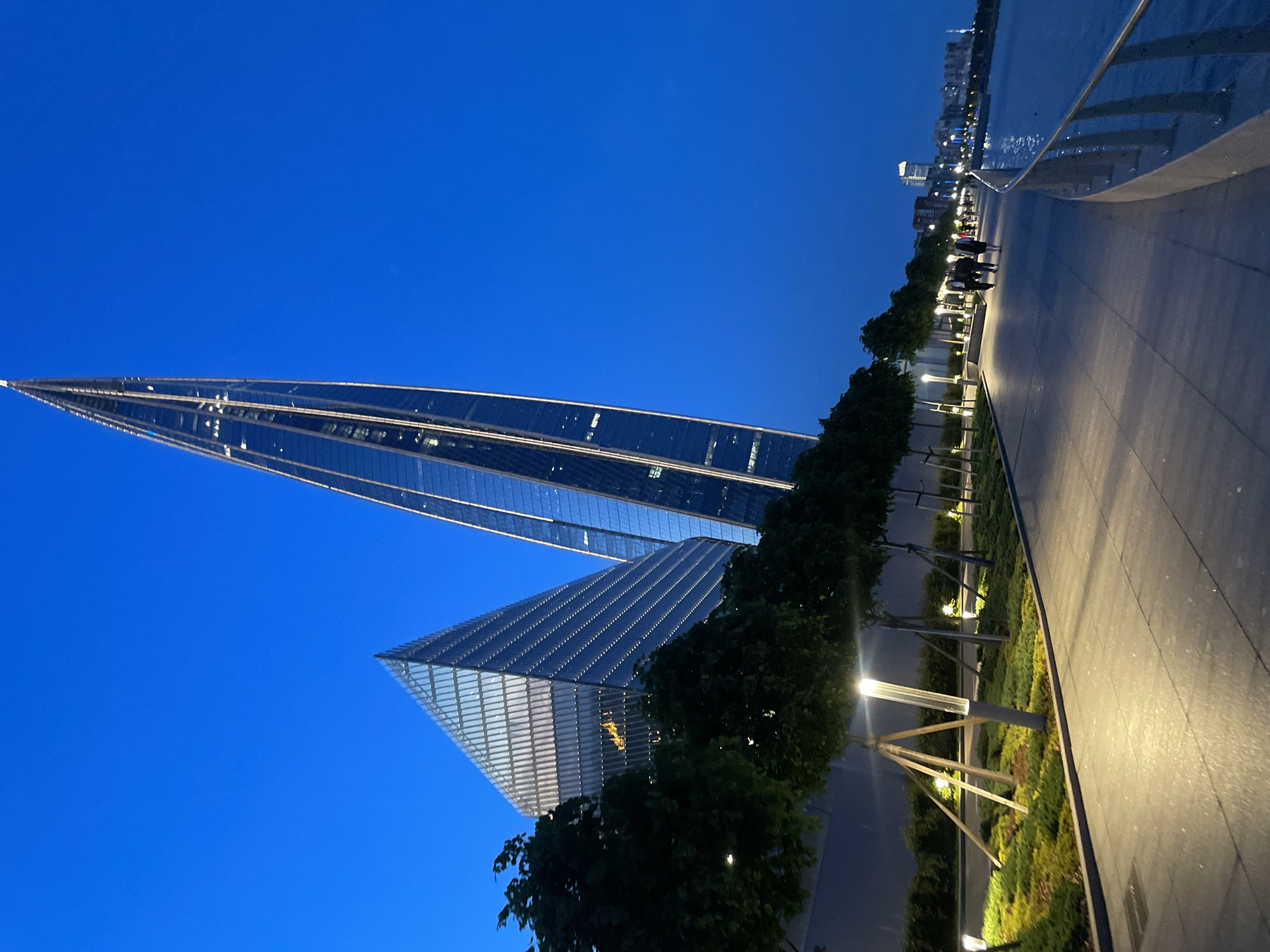 The image depicts the Lakhta Center in Saint Petersburg, Russia, at dusk. Two modern skyscrapers dominate the scene, with one a sharply pointed spire and the other a triangular prism. The sky is a deep twilight blue. In the foreground, a paved promenade runs alongside a body of water, with a metal railing bordering the water's edge. The promenade is lined with young trees staked with wooden supports and illuminated by modern, cylindrical lampposts casting warm light onto the path and surrounding greenery. Several people are walking along the promenade, some in pairs, their silhouettes visible against the illuminated path. In the background, other buildings can be seen along the waterfront, suggesting a developed urban area. The overall atmosphere is one of modern urban development at the close of day.
