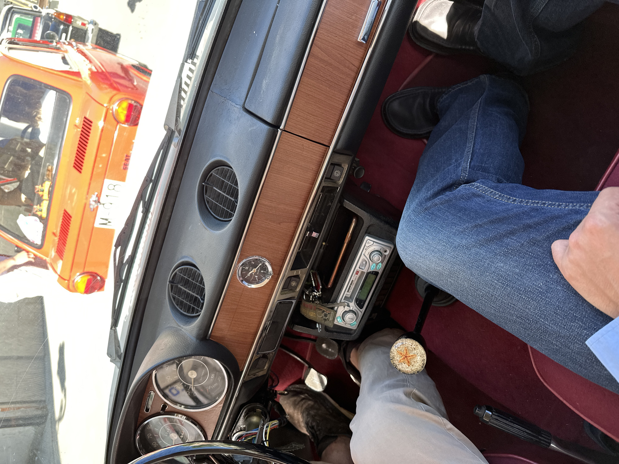 The image shows the interior of a classic car, likely taken from the driver's perspective. The dashboard is visible, featuring a wooden trim, air vents, gauges, a clock, and a modern-looking car radio. The driver's legs are visible, wearing blue jeans and black shoes. Another person's leg, wearing beige pants, is also visible near the gear stick. The gear stick has a decorative knob with a compass or star design.
Outside the car, through the windshield, a glimpse of an orange SEAT car and a license plate with the prefix "M" can be seen, which is indicative of Madrid, Spain. The license plate includes the numbers "618". The environment appears to be a street or parking area. The scene is captured in daylight.