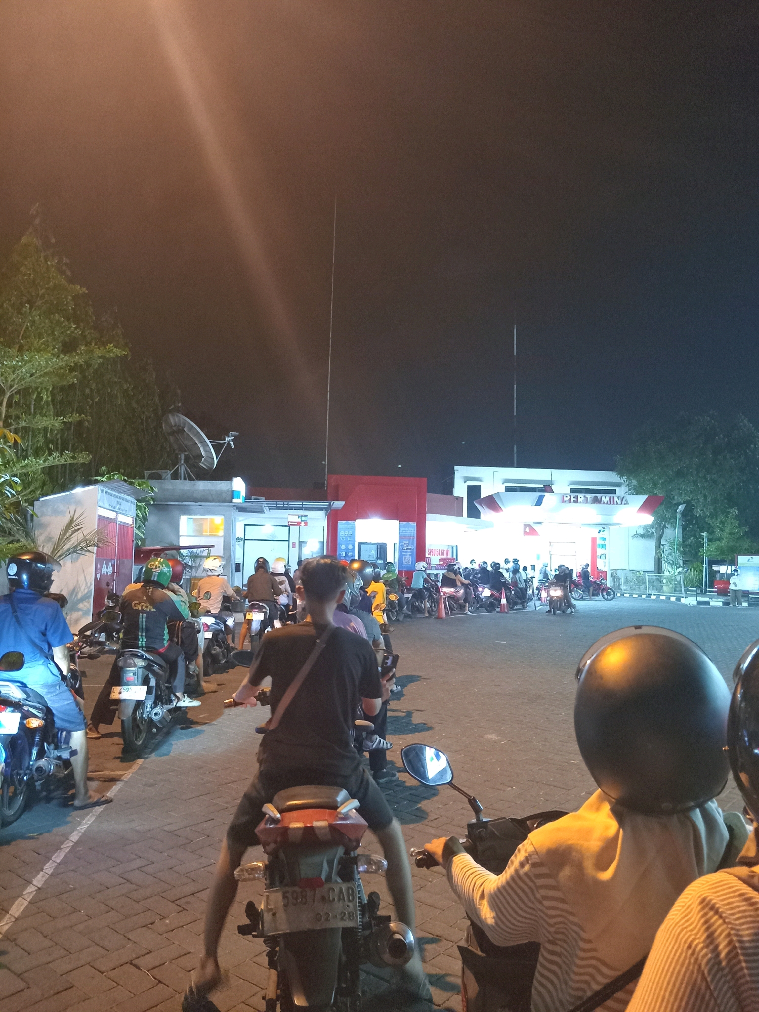 The image depicts a nighttime scene at a Pertamina gas station in Karangturi, Indonesia. A long queue of motorcycles and their riders are waiting. The gas station signage prominently displays "PERTAMINA". The ground is paved with interlocking bricks. Several people are seated on their motorcycles, facing away from the camera, with some wearing helmets. In the foreground, two individuals are on motorcycles, also facing away. To the left, a person wearing a blue shirt and shorts is on a motorcycle. Behind the queue, the gas station building with its illuminated pumps and canopy is visible. Trees are present on the left and right sides of the frame. A satellite dish is mounted on a structure behind the gas station. The lighting suggests it is nighttime, with artificial lights from the gas station illuminating the area.