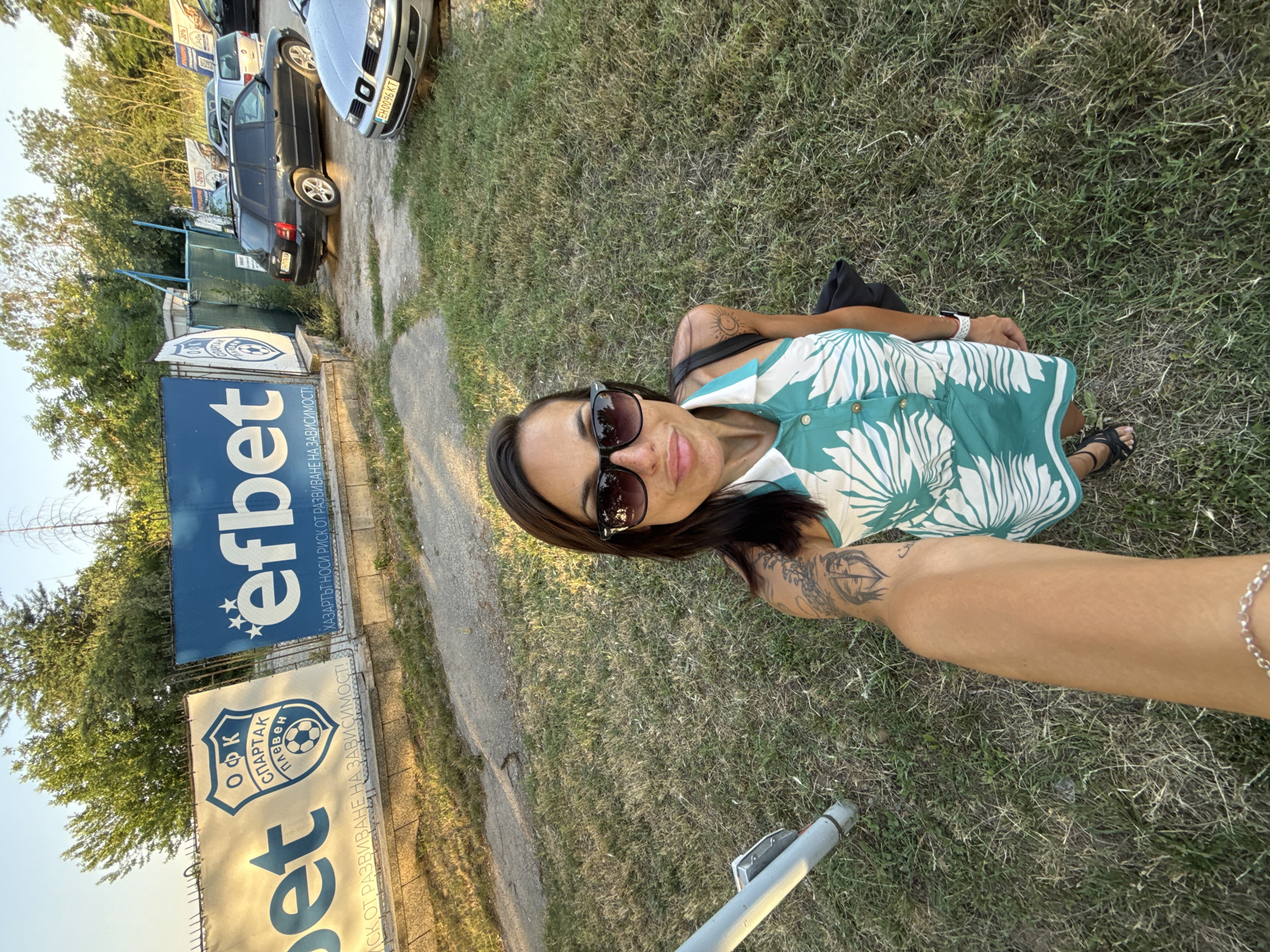 This media file captures a woman taking a selfie outdoors. She is wearing sunglasses and a teal and white patterned dress, with visible tattoos on her arm. She is lying on dry grass, with a black bag resting beside her. In the background, several cars are parked along a paved area. Adjacent to the parking area are large banners, one with the "efbet" logo and Bulgarian text, and another with the "OFK Spartak Pleven" crest and a soccer ball. The banners suggest the location is a sports venue, possibly a soccer field in Pleven, Bulgaria. The lighting indicates it's late afternoon or early evening, with the sun casting warm light and long shadows. The overall scene conveys a relaxed, casual atmosphere.