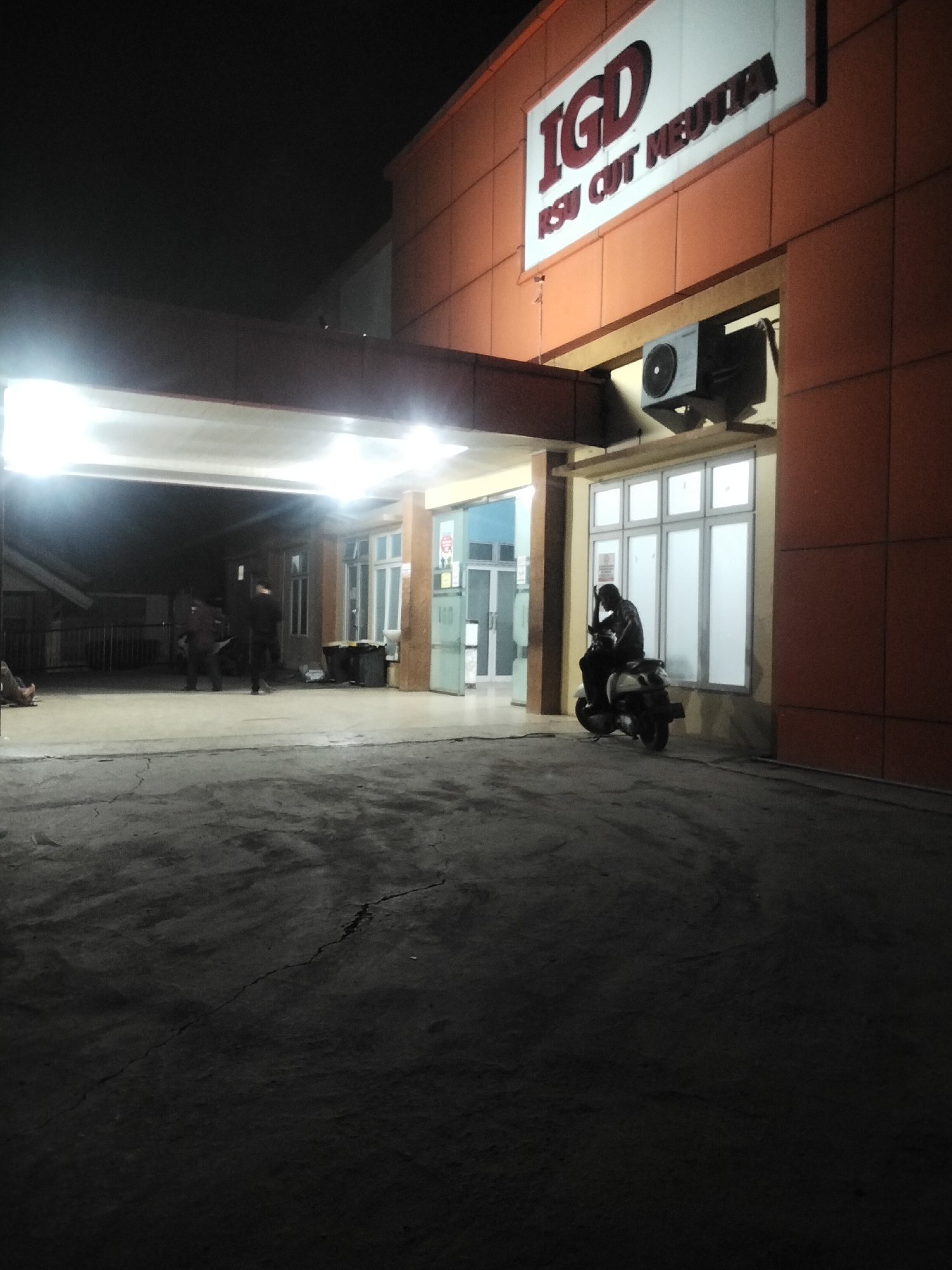 The image depicts the exterior of the IGD (Instalasi Gawat Darurat, or Emergency Room) at RSU Cut Meutia, a hospital located in Lhokseumawe, Indonesia. The building's facade is primarily orange-colored panels. A prominent sign above the entrance reads "IGD RSU CUT MEUTIA" in large, bold letters. An outdoor air conditioning unit is mounted on the wall below the sign.
In front of the building, several individuals are visible. One person is seated on a scooter, facing away from the camera, with their head bowed. To the left, two figures, possibly staff or visitors, are standing in the sheltered entrance area. The ground in the foreground is a dark, paved surface with visible cracks. The scene appears to be illuminated by artificial lights, suggesting it is nighttime. The overall atmosphere is quiet and dimly lit, typical of an emergency room entrance at night.