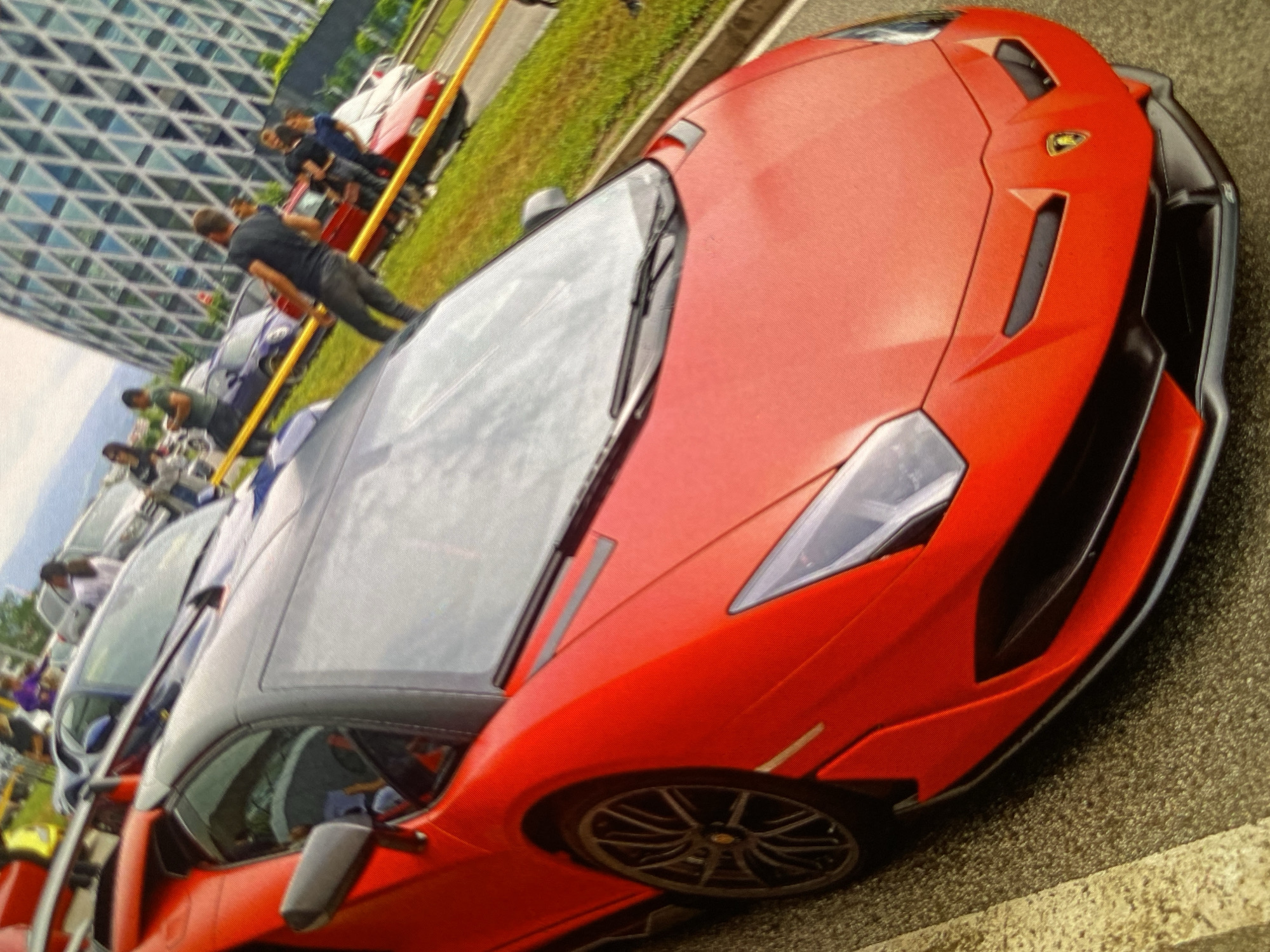 This image depicts an outdoor car gathering, likely a car show, in Bankya, Bulgaria, under clear daytime conditions. The primary subject is a striking, bright red Lamborghini sports car with a black roof and black aerodynamic details on its body. The iconic golden bull emblem of Lamborghini is clearly visible on its front hood.
Numerous people are present, observing the vehicles. In the mid-ground, one person wearing a black t-shirt and dark pants leans on a yellow barrier or pole, appearing to watch the event unfold. Other individuals are scattered throughout the background, some near other parked cars.
Beyond the featured red Lamborghini, several other vehicles are visible, contributing to the car show atmosphere. These include a white or silver modern car, a blue car, and a classic-style red car further back. The setting includes an asphalt road surface and a grassy verge.
A prominent architectural element in the background is a large, modern building with a distinctive geometric, grid-patterned glass facade that reflects the bright sky. The overall scene conveys an atmosphere of interest and appreciation for high-performance automobiles. Aside from the Lamborghini emblem, no other text is discernible.