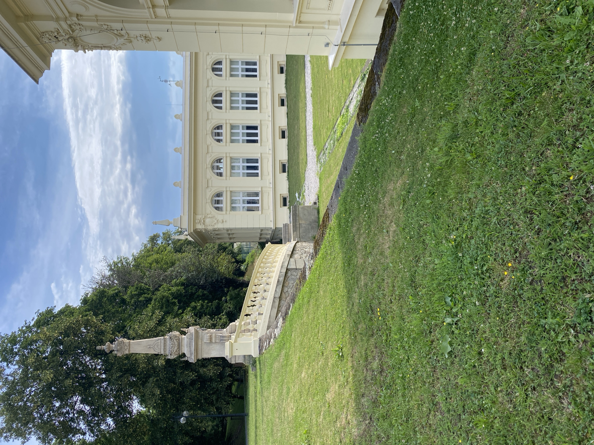 The image shows a view of the Bílina Castle in Bílina, Czechia. In the foreground, a lush green lawn slopes upwards towards a cream-colored stone balustrade, which curves to the right. Beyond the balustrade, a monument stands tall, topped with an ornate finial. Behind this, a large, light yellow building with arched windows and decorative elements is visible. Further in the background, dense green trees form a backdrop, with a blue sky dotted with white clouds overhead. The scene is quiet and serene, with no people present.