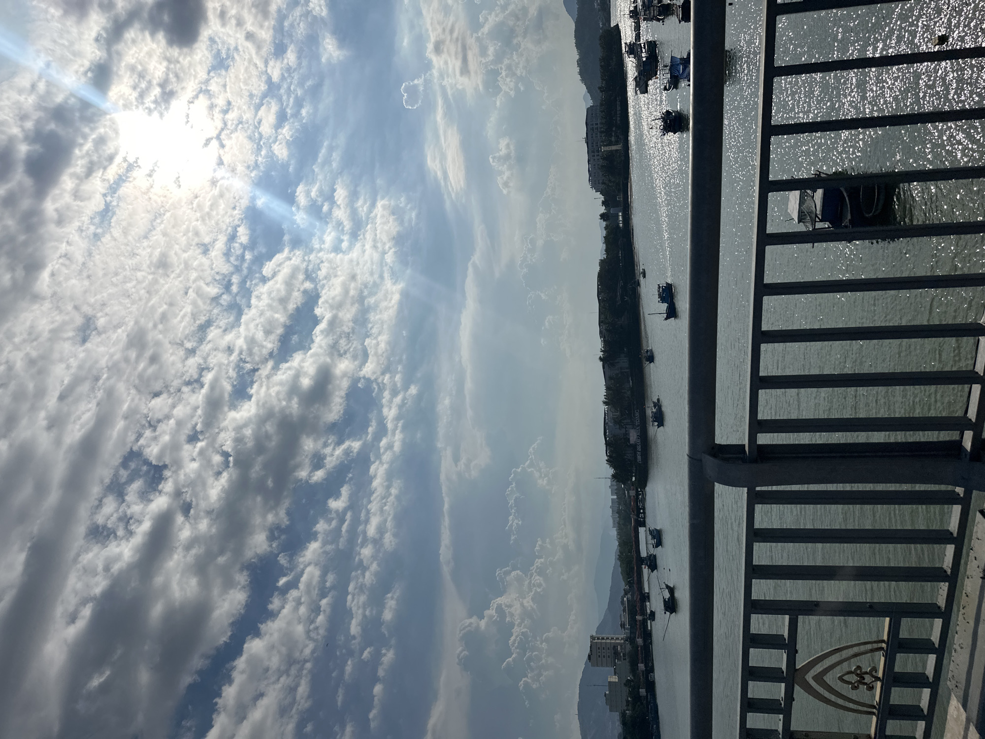 The image presents a view from a bridge, likely in Bac Nha Trang Ward, Vietnam, based on visual cues. The sky is filled with clouds, and the sun is positioned near the top of the frame. Below the sky is a body of water, possibly a river or bay, with a shimmering surface reflecting the sunlight. Several boats are visible on the water. A cityscape is on the shoreline along the water, with buildings visible. A bridge railing is in the foreground, partly obscuring the water and cityscape. The image is slightly rotated.