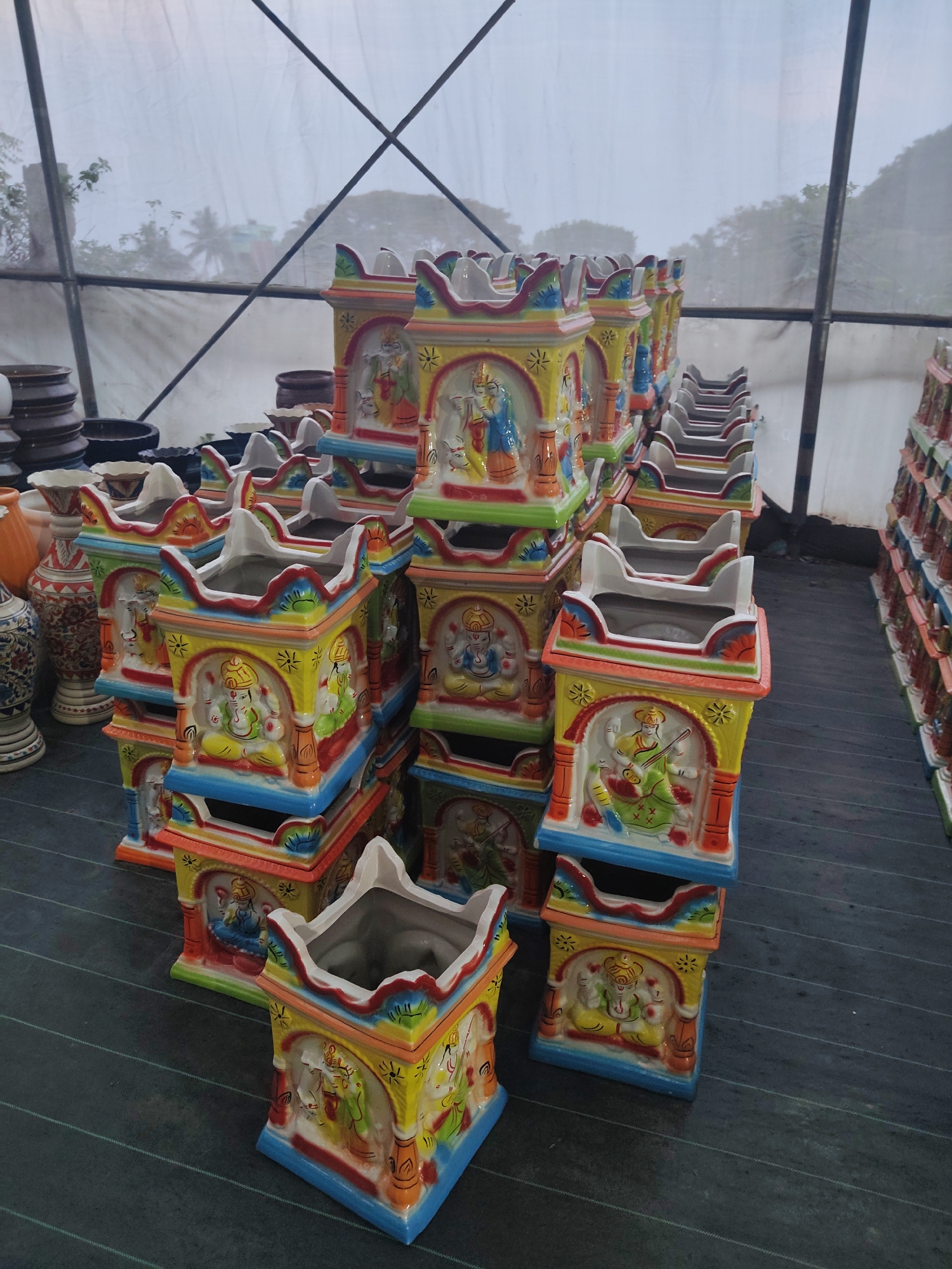 The image shows a large collection of colorful, hand-painted ceramic pots stacked in rows. The pots are square-shaped with decorative edges and feature images of Hindu deities. The location appears to be a greenhouse or covered market, indicated by the translucent roof and metal support structure visible in the background. The pots are arranged on a dark, possibly paved, surface. It is inferred that the location is ಚಂದ್ರಪ್ಪ ವೃತ್ತ, India due to the images metadata tags, which describe this location and the artifacts.