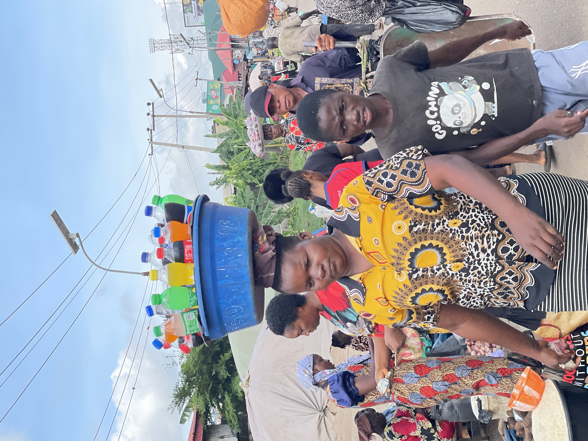 This image captures a bustling outdoor market scene in Unknown, Unknown. In the foreground, a woman walks towards the viewer, balancing a large blue basin filled with assorted plastic bottles of beverages (including sodas in red, green, yellow, orange, and clear bottles) on her head. She wears a yellow and black patterned short-sleeved top featuring floral and animal print motifs, over a black and white striped skirt. To her immediate right and slightly behind, a young boy walks, wearing a dark grey t-shirt depicting a panda character with a blue scarf and the text "GO! CHINA." He wears light blue shorts.
Behind these two figures, the market extends with various individuals and stalls. Another woman in the mid-ground, to the left, is also seen carrying an item on her head, wearing a red and white floral patterned top. Further left, a woman in a blue headscarf and a colorful patterned dress appears to be engaging in a transaction, holding an item and possibly money, next to a large white market tent. On the far left, a black bucket containing white grains (possibly rice) and an orange plastic bowl are visible.
In the mid-ground, to the right of the foreground woman, a man wearing a black baseball cap and a dark shirt holds a metal pole. Behind him, several other people are present, some carrying goods on their heads or engaged in market activities. The background features more market stalls, some with green awnings, as well as dense green foliage, likely banana trees. Utility poles with numerous power lines crisscross the sky, which is overcast with scattered clouds. A street light with a solar panel is also visible. Further in the background, a large advertising billboard with "Pepsodent" branding and another sign indicating "200" can be seen, alongside a metal telecommunications tower structure. The ground appears to be a mix of dirt and asphalt.