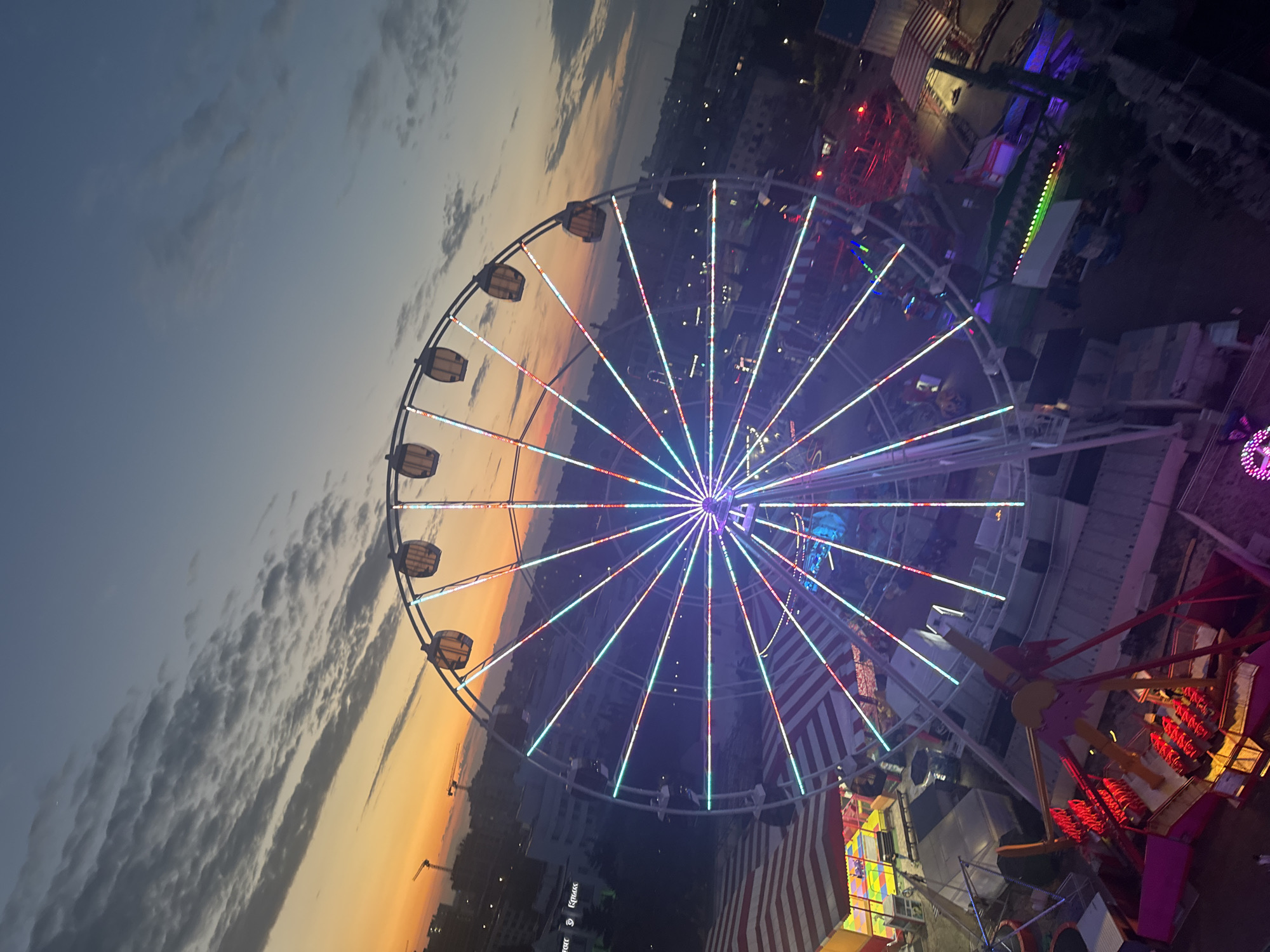 An eye-level, dusk-lit photograph shows a large Ferris wheel at a fair in Yevpatoriya, Ukraine. The Ferris wheel is lit with colorful LED lights forming lines of red, white, and blue across the center of the wheel. The sky is a blend of orange sunset to the left and a fading blue with wispy clouds to the upper left. A few buildings can be seen on the distant horizon. To the right of the Ferris wheel are various fair rides and carnival attractions with colorful lights. An American flag is visible in the foreground.