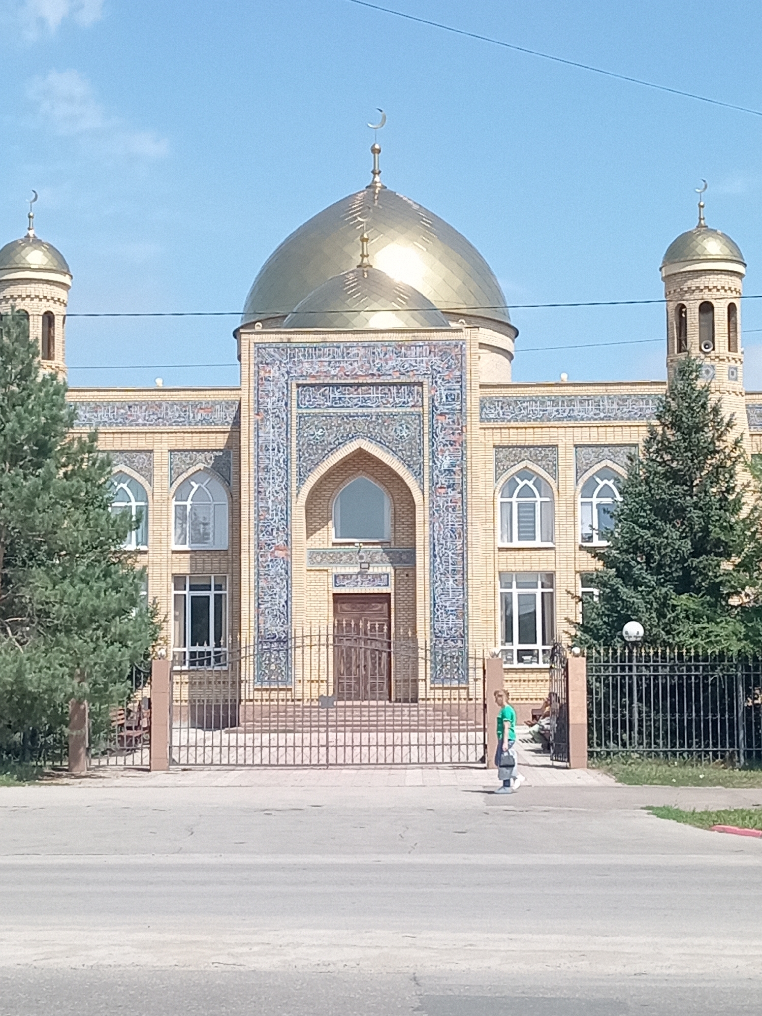 A wide-angle shot captures the exterior of a mosque with two minarets and a large golden dome against a clear blue sky. The mosque's facade is constructed from light-colored bricks and adorned with intricate blue and white mosaic tiles arranged in decorative patterns and Arabic calligraphy. A large arched entryway is visible in the center, flanked by tall windows with arched tops. A black metal gate and fence enclose the mosque grounds. In front of the fence, a paved pathway leads to the entrance, and beyond that, a road runs across the foreground. A person in a green t-shirt and light-colored pants walks across the paved area to the right of the entrance, carrying a bag. Two evergreen trees are positioned on either side of the mosque, partially obscuring its facade. This image is of the mosque in Rudny, Kazakhstan.