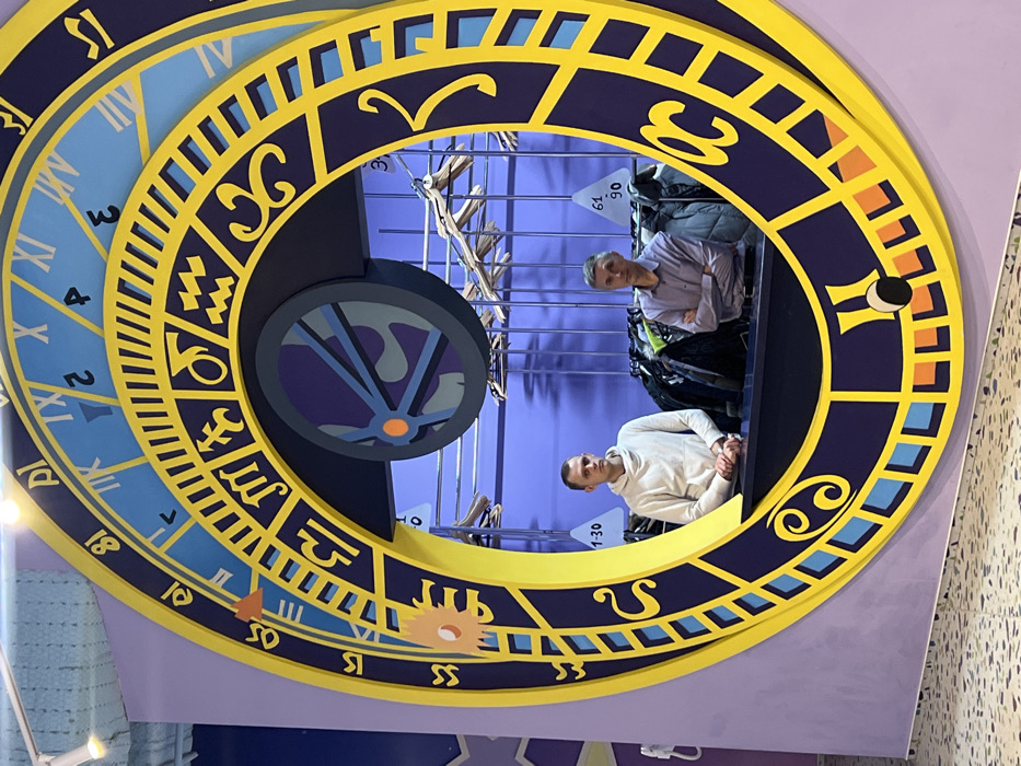 The image depicts two men standing behind a large, circular clock-themed structure. The clock features Roman numerals and zodiac symbols in shades of blue, purple, and yellow. A smaller wheel with blue and gray patterns is attached to the side of the clock. Clothing hangs on racks in the background behind the men. The image is angled, providing a unique perspective of the scene.