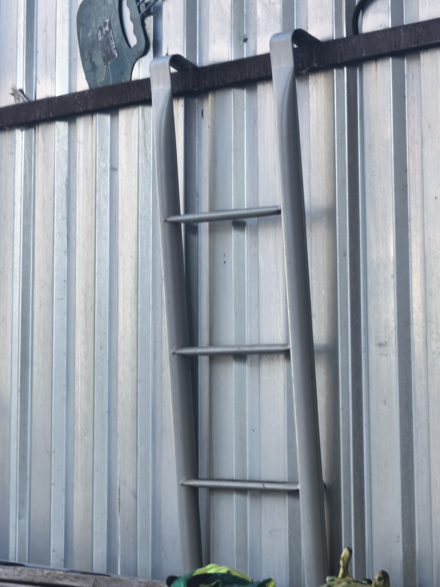 An outdoor shot shows a close-up of a silver-colored ladder with three rungs against a corrugated metal wall. The ladder is positioned vertically, and its top is obscured by a dark brown horizontal beam. A green object, possibly a tool or bag, is visible on the beam to the left of the ladder. The corrugated metal wall has a metallic sheen and vertical ridges. The lighting suggests it is daytime, with some shadows cast on the wall.