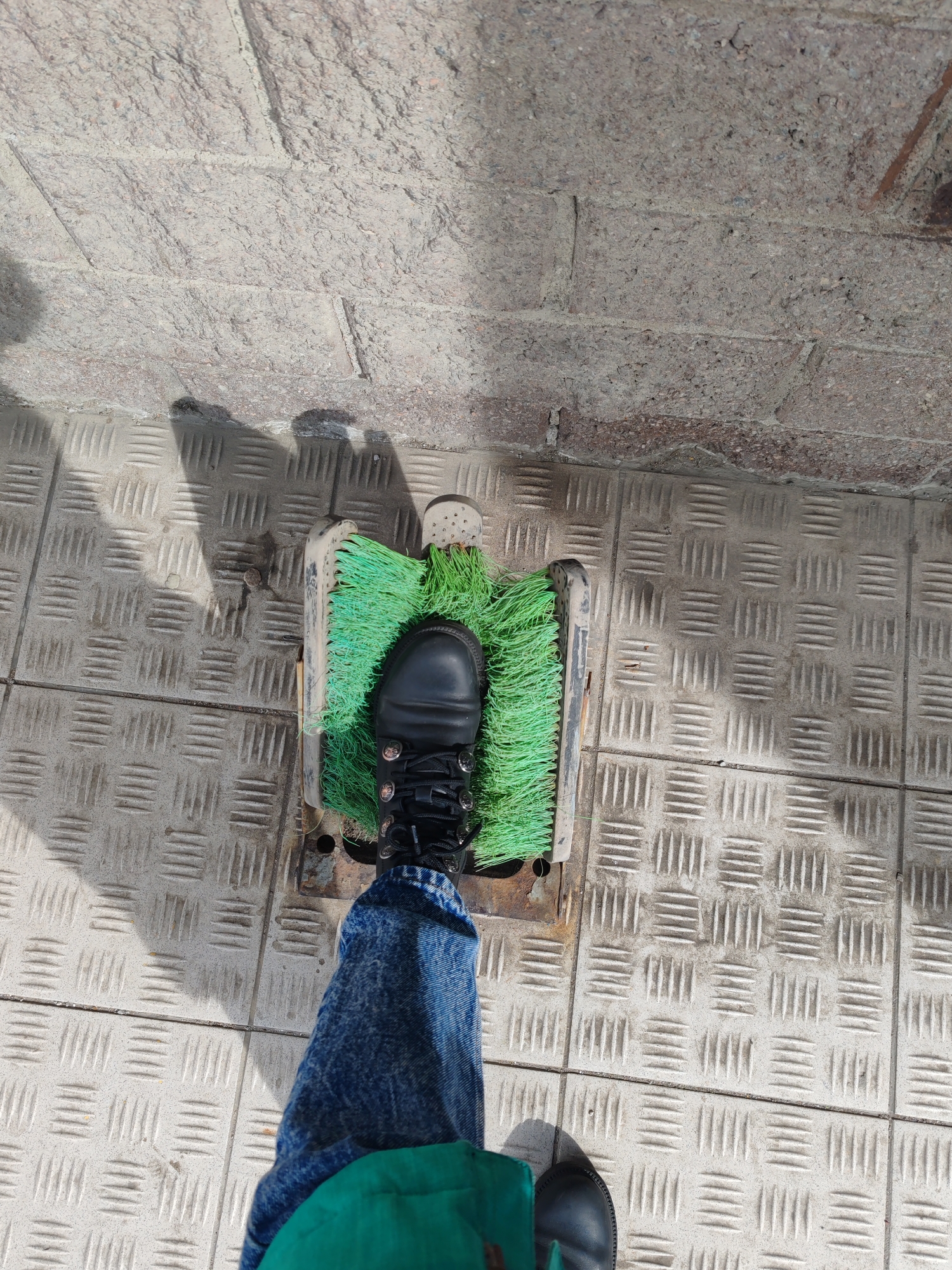 The image presents a high-angle, overhead view of a person's legs and feet, standing outdoors. One foot, encased in a black combat-style boot with silver eyelets and black laces, is placed firmly on a boot cleaner. The boot cleaner features vibrant green synthetic bristles, suggesting it is well-used but still functional. The other leg is clad in blue denim jeans, with a portion of a green garment (possibly a jacket or bag) visible near the foot, which is partially out of frame on the patterned ground.
The ground surface consists of two distinct textures. In the foreground and extending to the left, there is a gray metal grating or plate with a raised, non-slip diamond-plate pattern. Adjacent to this, in the background and extending to the right, is a rough, coarse-textured concrete or stone surface, possibly a wall or uneven paving.
Strong, defined shadows are cast across the patterned metal surface, indicating bright sunlight and a clear sky, likely during the daytime. The scene suggests a momentary pause at an entrance or outdoor area where footwear might need cleaning. There is no visible text in the image. The location context provided is Saint Petersburg, Russia.