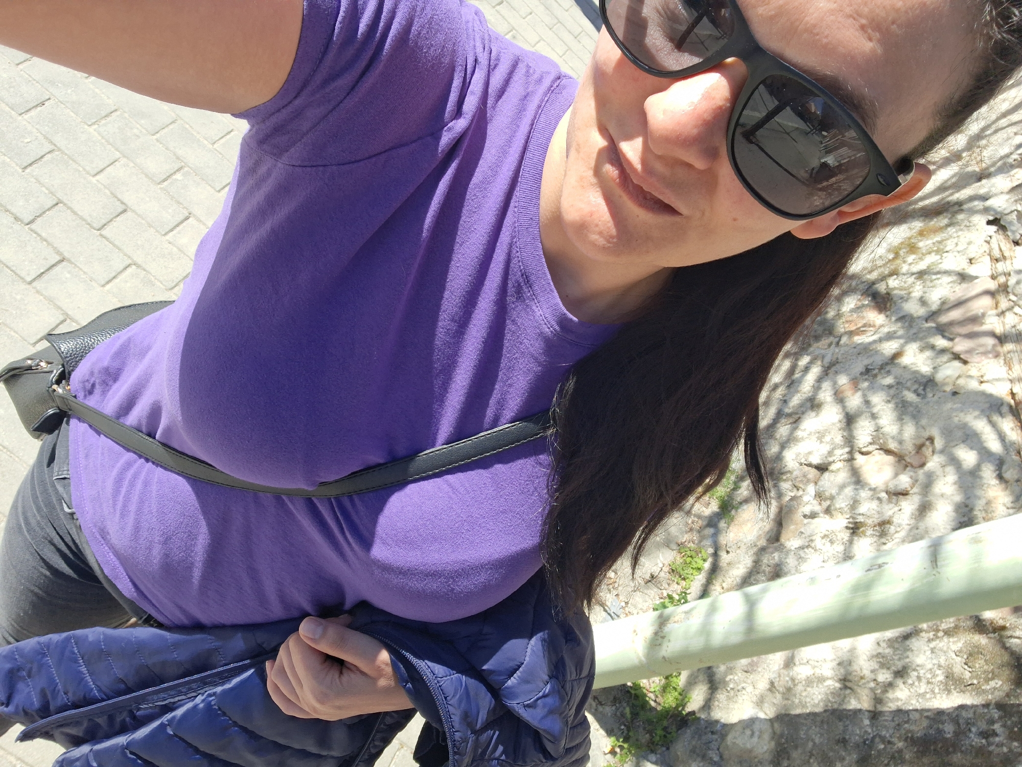 A woman wearing a purple t-shirt and sunglasses stands outside in Goce Delcev, Bulgaria. A black crossbody bag is visible on her left side. She holds a blue puffer jacket in her left hand. The ground is paved with light-colored bricks on the left and rocky terrain with sparse vegetation on the right. A light-colored metal railing runs along the right side of the frame.