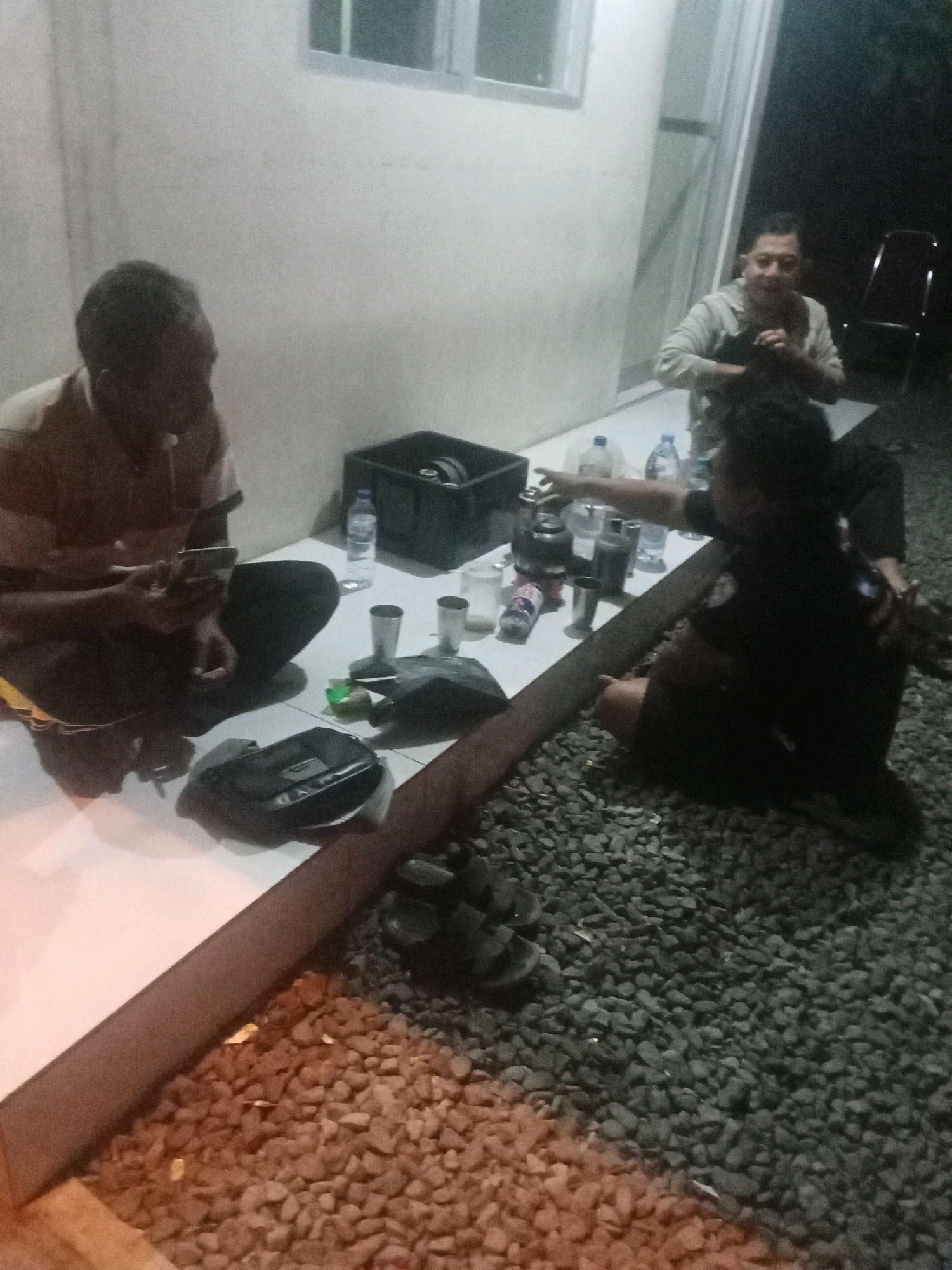 Three individuals are seated on the ground near a building in Jombang, Indonesia. One person in the foreground is looking at a smartphone. In the middle ground, another person is leaning forward, appearing to interact with something on a low table, possibly pointing. In the background, a third person is seated, looking toward the other two. Several water bottles, cups, and a black container are visible on the table. The ground is covered with gravel. The setting appears to be outdoors, adjacent to a building with windows.
