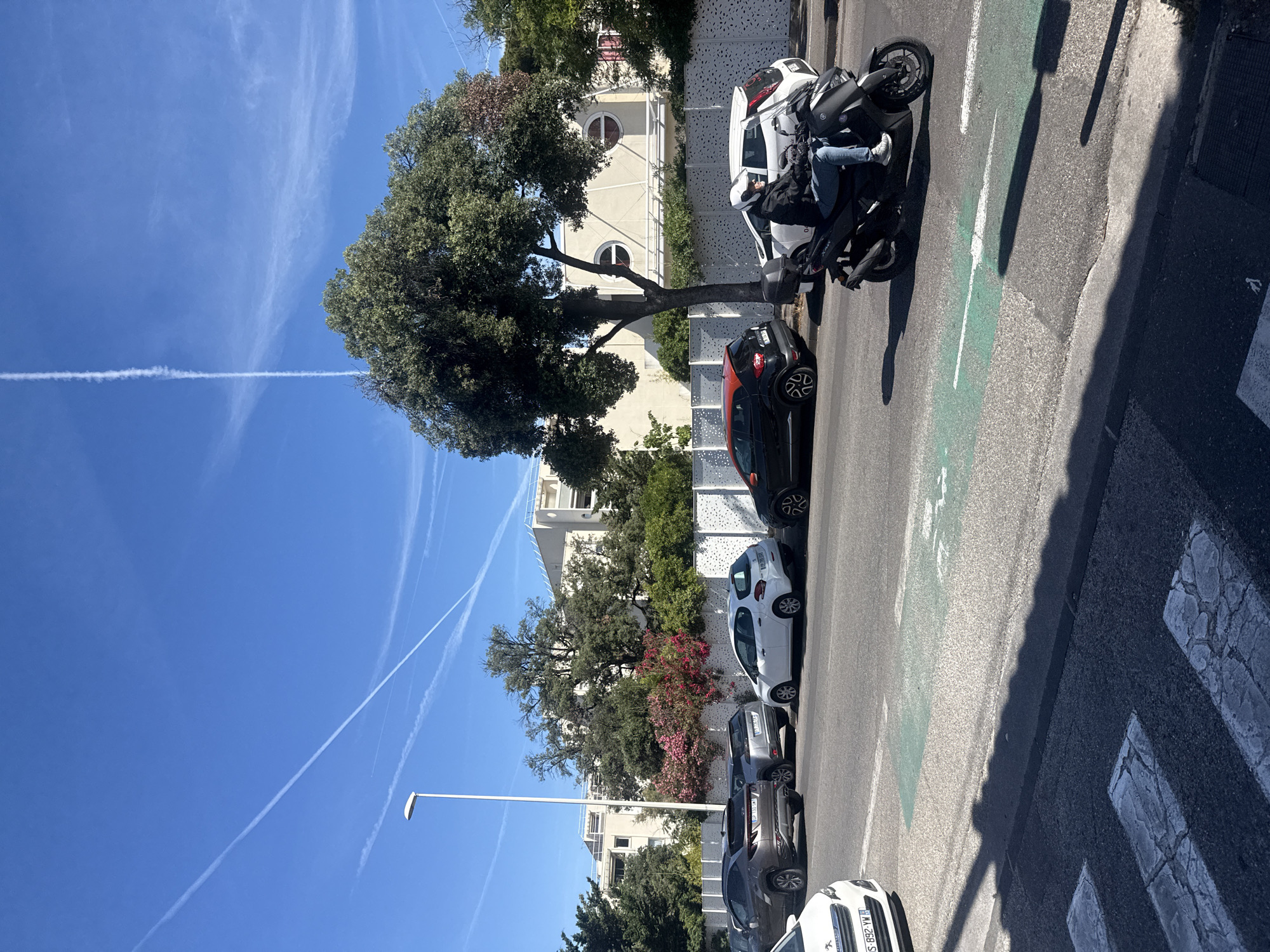 The image depicts a street scene with several cars parked along the roadside. A person is riding a black scooter, wearing a helmet and blue jeans. The scooter is on the right side of the image, moving towards the viewer. There are trees and buildings in the background, with a clear blue sky overhead featuring contrails from airplanes. The street has a pedestrian crossing in the foreground and a designated bike lane marked with green paint. The overall environment appears to be an urban setting on a sunny day.
