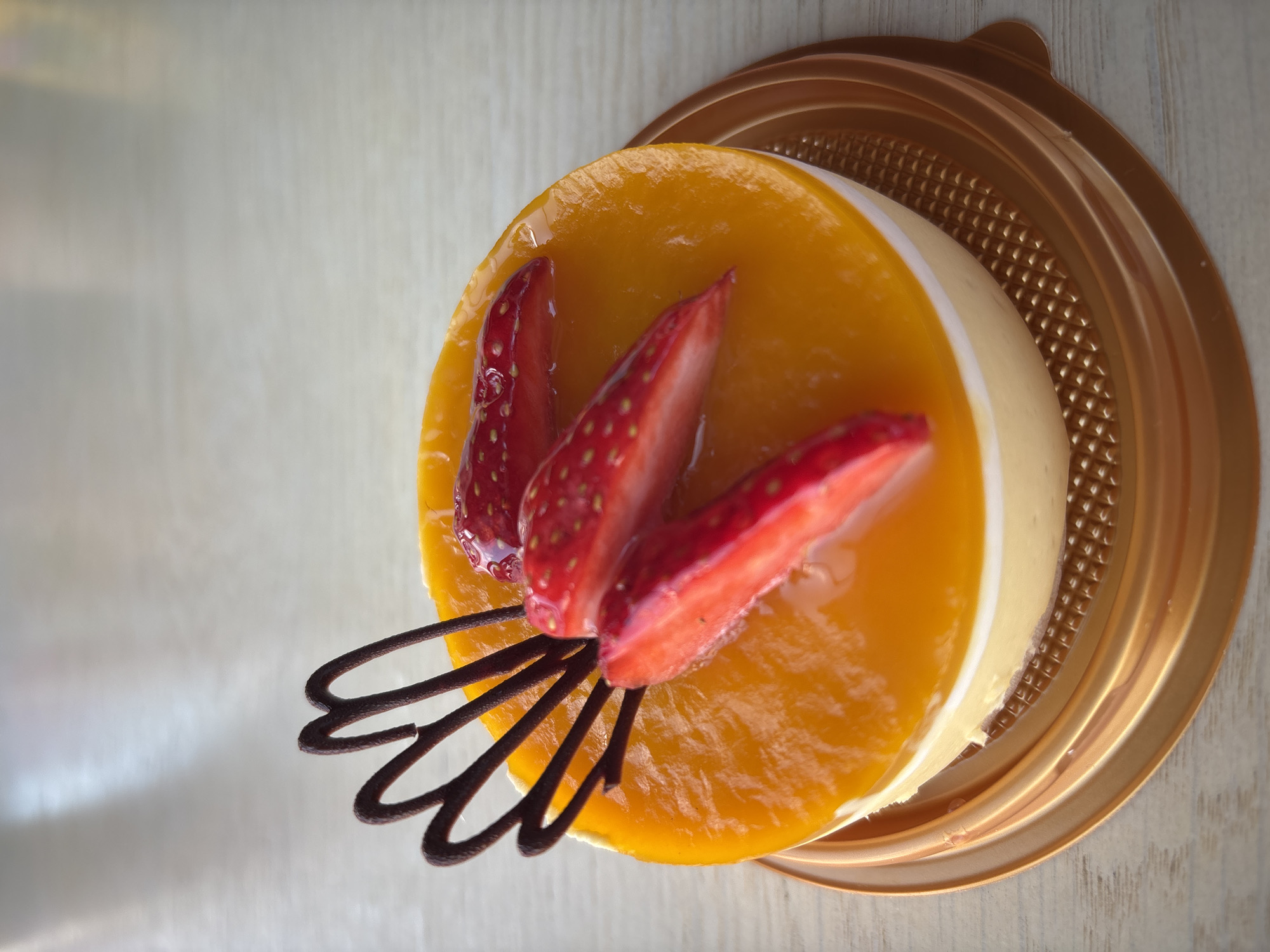 A top-down, close-up shot reveals a round dessert on a gold-colored plastic plate. The dessert features a yellowish-orange glaze. It is garnished with three sliced strawberries and a dark-brown, chocolate-like decoration. The dessert sits on a light-colored wooden surface. There are no people in the image. The location is not possible to determine from the image.