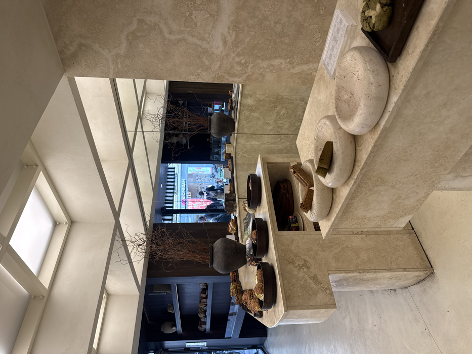 The image depicts the interior of Tapisserie Patisserie, located at 6 Rue Sainte-Croix de la Bretonnerie, 75004 Paris, France. The scene is a minimalist, modern retail space characterized by light-colored, textured stone surfaces, likely travertine, forming the display counters and a partial wall on the right.
The main display features an L-shaped counter presenting a variety of baked goods and pastries in dark ceramic bowls and on platters. Items visible include various sizes and shapes of bread and pastry products. On the front-most counter section, three rounded, light-colored stone bowls, one containing a light-brown baked item and another a green-tinted item, are visible next to a small wooden board holding a green-speckled cookie.
Two large, dark pottery vases containing tall, dry, brown branches serve as decorative elements on the main display counter. The ceiling features a grid of illuminated panels and a central square skylight, providing overhead lighting.
A large mirror occupies the upper section of the back wall, reflecting the street scene outside the establishment. The reflection shows several blurred figures, likely pedestrians, and a storefront with a black and white striped awning and the reversed text "MADEMOISELLE." To the left, a dark wooden shelving unit displays additional baked items and pottery. The floor is a light, smooth concrete. The overall environment is clean, well-lit, and uncluttered.