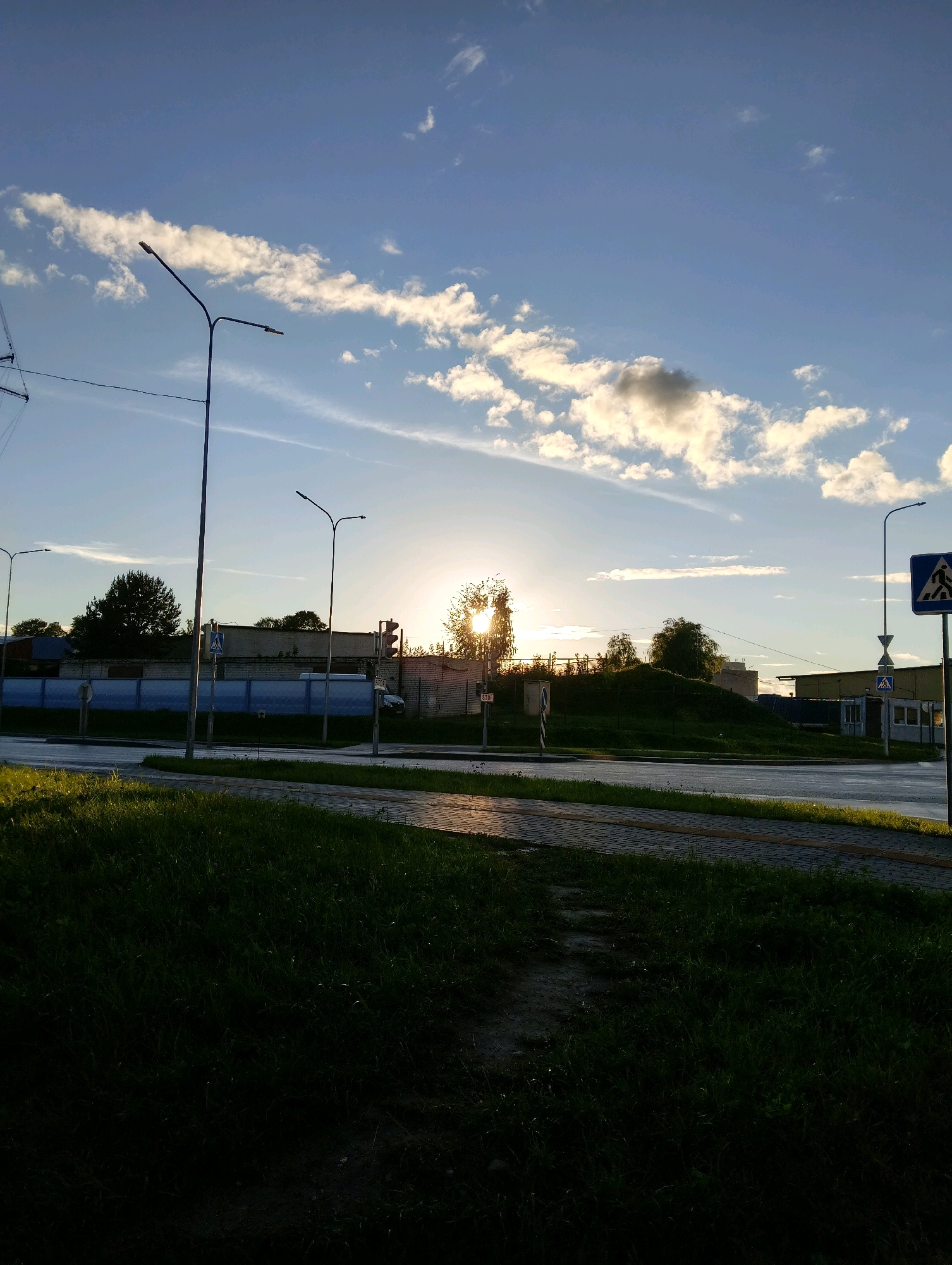 The image depicts a street scene in Hrodna, Belarus, during sunset. The sky is a vivid blue with scattered white and gray clouds, illuminated by the setting sun which is partially obscured by trees in the middle ground. The sun's rays create a bright glare, casting long shadows.
In the foreground, lush green grass lines a paved walkway and a road. The road appears wet, possibly from recent rain, reflecting the light. On the left side of the road, a tall street lamp stands prominently. Across the road, a low-rise building with a light-colored facade is visible, behind which are more trees and a hill. Another street lamp is positioned on the right side of the road, near a pedestrian crossing sign. A traffic light is also visible on the right, showing a red signal. Several other street lights and utility poles are scattered throughout the scene. The overall impression is that of an urban environment transitioning into evening.