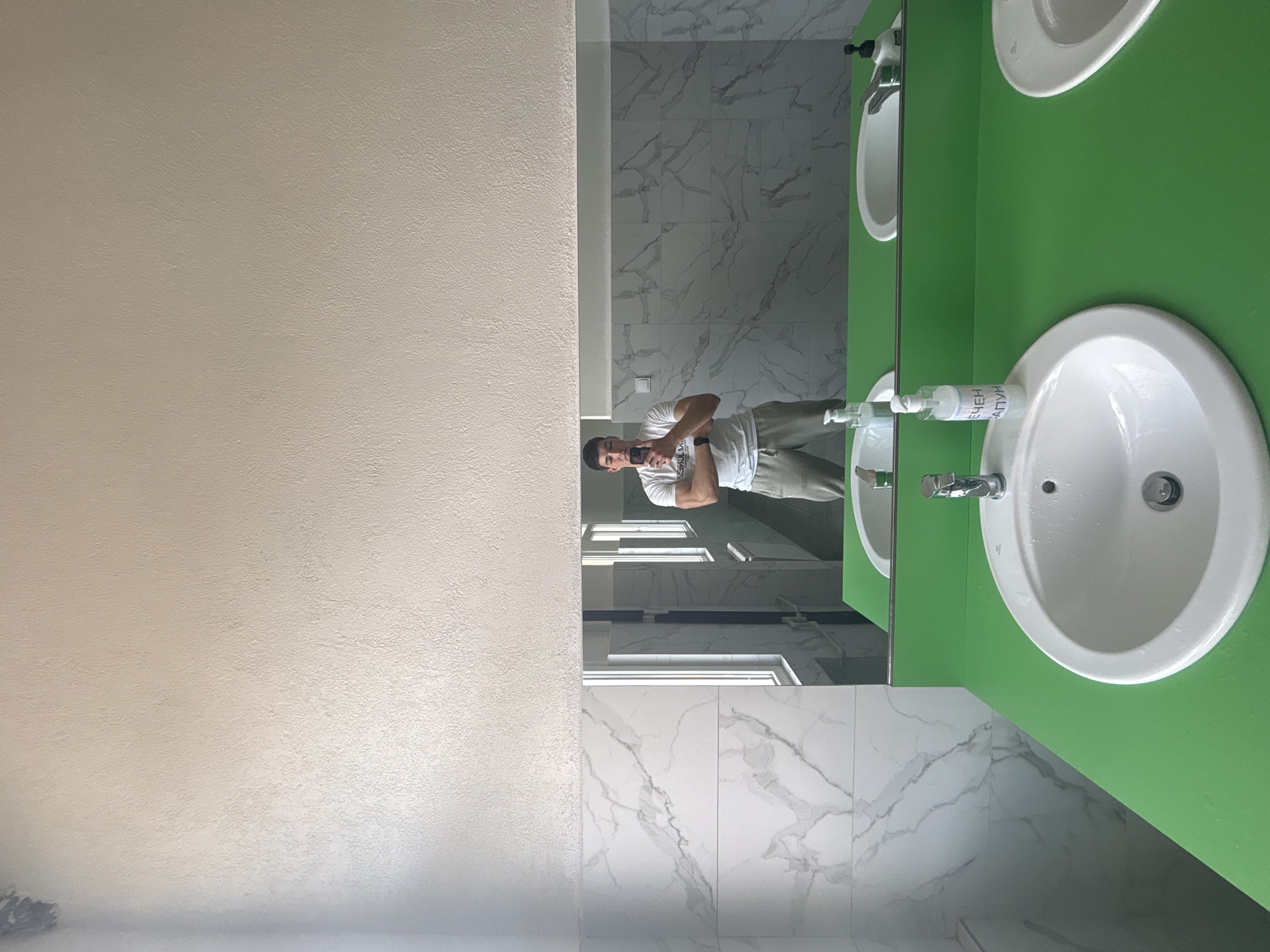 A man is captured in a mirror selfie within a modern bathroom or public restroom in Zlatograd, Bulgaria. The man, reflected in the mirror, is positioned centrally, facing the camera with his arms crossed, holding a smartphone. He is wearing a white t-shirt and light-colored trousers.
The setting consists of a large mirror on the right side of the image, reflecting a wall with white marble-patterned tiles and what appear to be windows above the man's head, indicating daytime. To the left, a textured, light beige wall fills a significant portion of the frame. Below the mirror, a vibrant lime green countertop features two white oval sinks, each with a chrome faucet. Between the two sinks, a clear pump bottle, likely containing soap or sanitizer, stands on the green surface.
Visible text on the pump bottle, which appears reversed due to the mirror reflection, reads "ЕН НАЧ" on its upper line and "АЛУН" on its lower line. The overall scene is well-lit and conveys a sense of cleanliness and contemporary design. The man's expression is neutral, suggesting a casual photo opportunity.