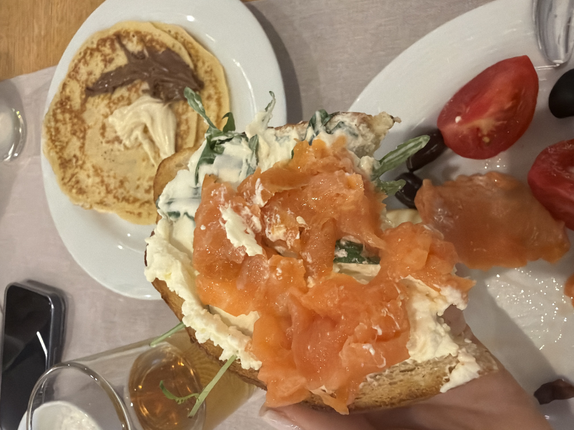 The image shows a close-up of a meal. In the foreground, a person's hand is holding a slice of toasted bread topped with cream cheese, salmon, and greens. Behind the toast, a plate holds several pancakes with dollops of butter and chocolate-hazelnut spread. Further back, a glass of amber liquid, possibly juice or beer, and a smartphone are visible. A white plate with a sliced tomato and black olives is also present in the foreground. The scene appears to be set on a light-colored tablecloth, suggesting a dining environment. The location is not specified in the image.