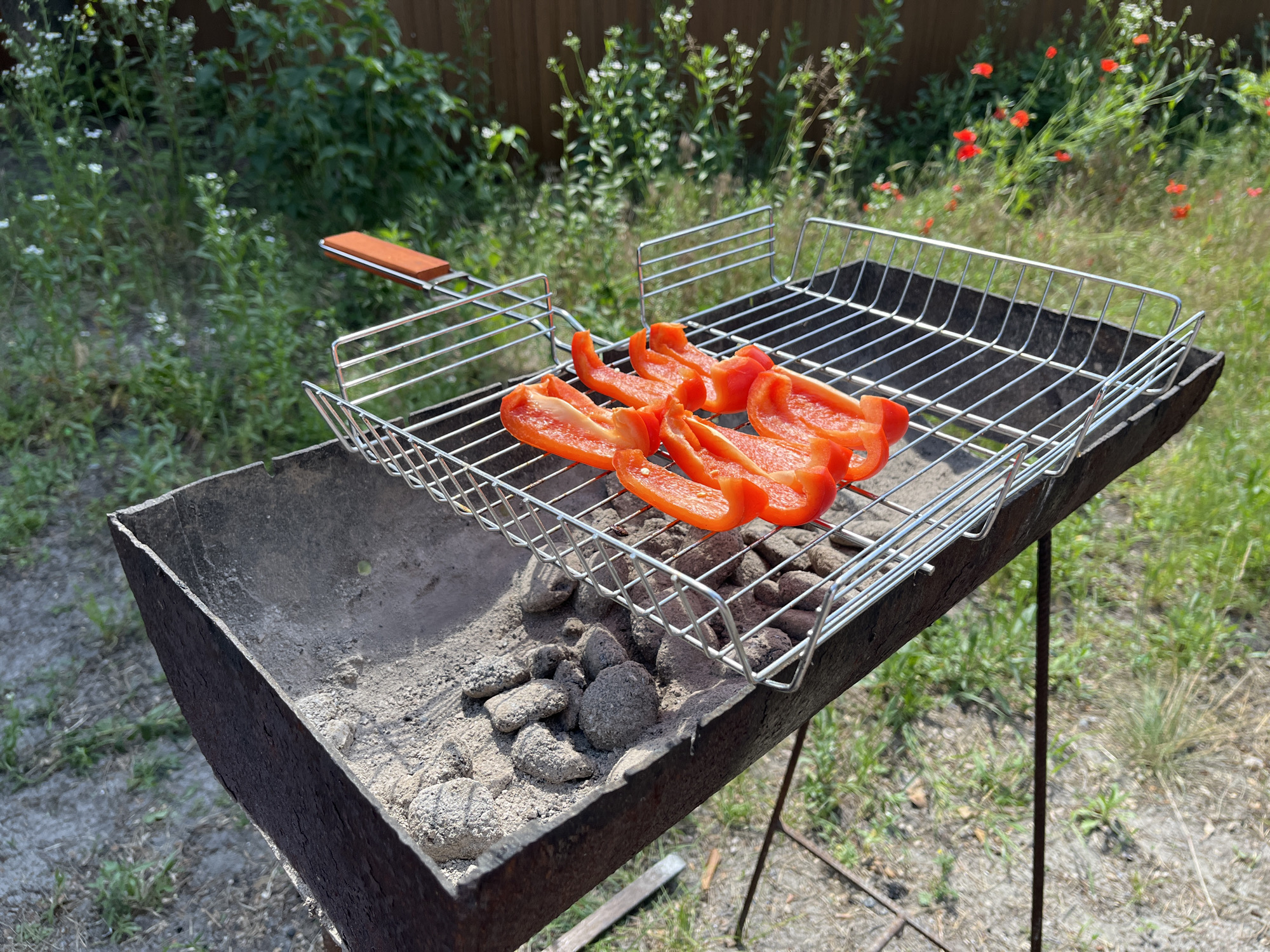A close-up, outdoor, eye-level shot shows a metal grill with a wooden handle positioned over a charcoal barbecue grill. Four bright red pepper slices are arranged on the top grill rack. The barbecue grill contains grey ash and charcoal briquettes. The background features lush green foliage, a wooden fence, and red wildflowers, suggesting a natural, possibly rural or suburban, environment. The lighting indicates it is daytime.