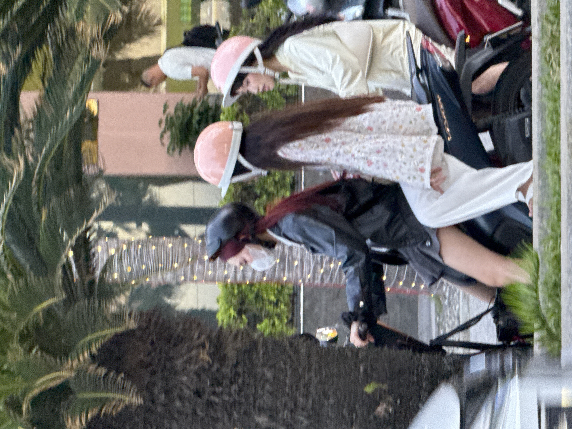 Three young women ride on a scooter in Nha Trang Ward, Vietnam. The woman in front is wearing a black jacket, a gray skirt, and a white face mask. She has long, dark red hair and is wearing a black helmet. The middle woman is wearing a white floral print top and white pants. She has long, dark hair and is wearing a light pink helmet. The woman in the back is wearing a light-colored long-sleeved top and is also wearing a light pink helmet. The scooter is dark blue. They are riding on a street with palm trees and a building in the background. A man in a white shirt and black backpack is visible in the distance. The lighting suggests it is daytime, possibly late afternoon or early evening.