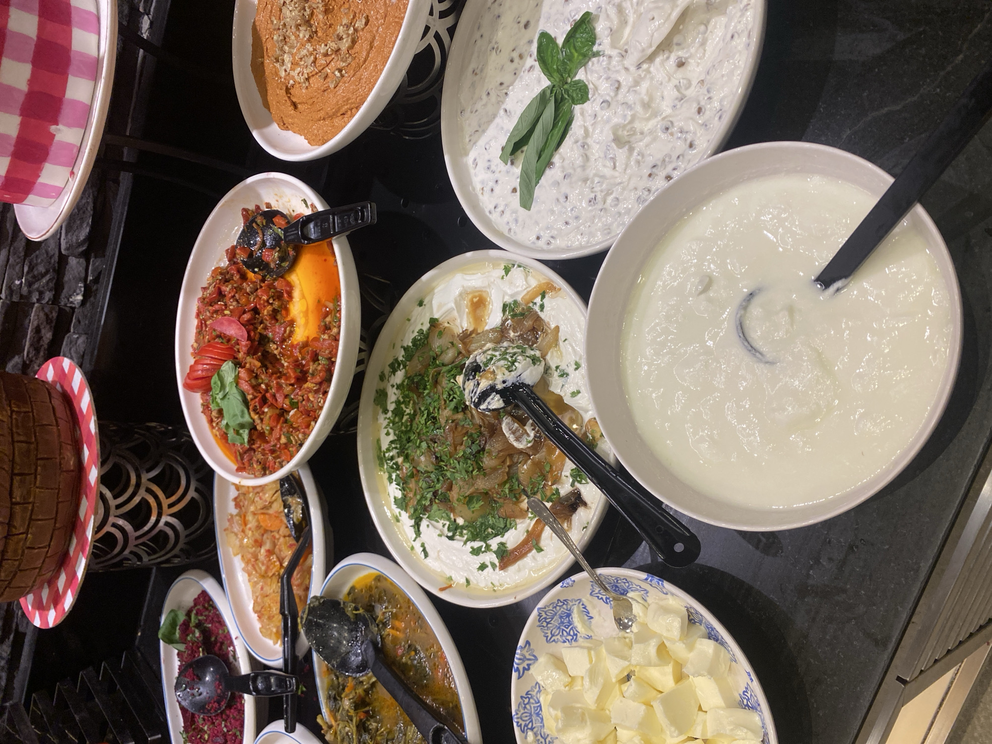 The image shows a buffet table with various food dishes. In the foreground, there is a bowl of white yogurt with a black serving spoon. To the right is a bowl of a creamy white dip with dark flecks and green basil leaves. To the left of the yogurt bowl, a white bowl with a blue pattern is filled with cubed white cheese. Behind these bowls are several other dishes, including a salad with chopped red ingredients, a dish with leafy green vegetables in broth, a bowl of rice or grains, and a vibrant red dish with tomatoes and herbs. In the background, there is a tiered cake stand with a cake decorated with pink and white checks, and a brown cake resembling a brick structure. The buffet is set on a dark countertop. The environment appears to be an indoor restaurant or catering setup in Aksu, Turkey. There are no people visible in the image.