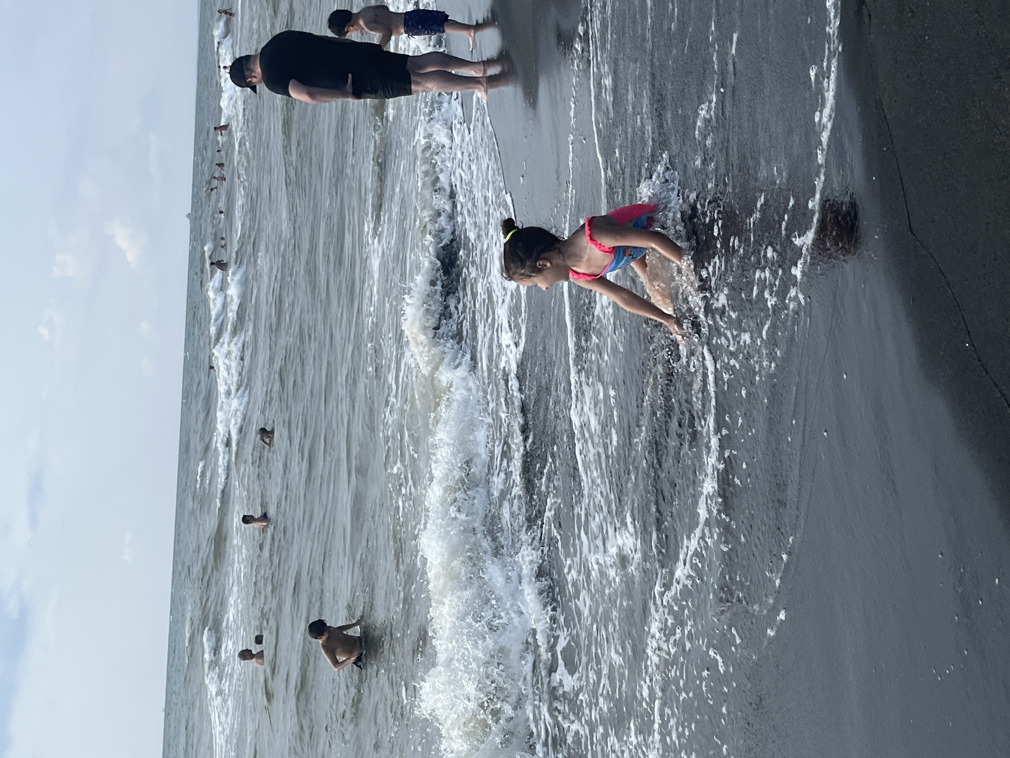 This media file captures a beach scene with several people enjoying the water. The primary subjects are a group of individuals, including adults and children, in and around the sea. A young girl in a pink and blue swimsuit is prominently featured in the foreground, interacting with the waves. In the background, several people are visible wading or swimming in the water. The scene is set on a sandy beach with the sea in the background.
The activity taking place is leisure and recreation at the beach. People appear to be swimming, playing in the water, and enjoying a day out. The weather appears to be overcast, with a calm sea and gentle waves. The time of day is not explicitly clear but seems to be daytime. There are no visible objects other than people and the natural elements of the beach and sea. No text is visible in the media file. The location context provided is Ureki, Georgia, which is known for its magnetic black sand beaches.