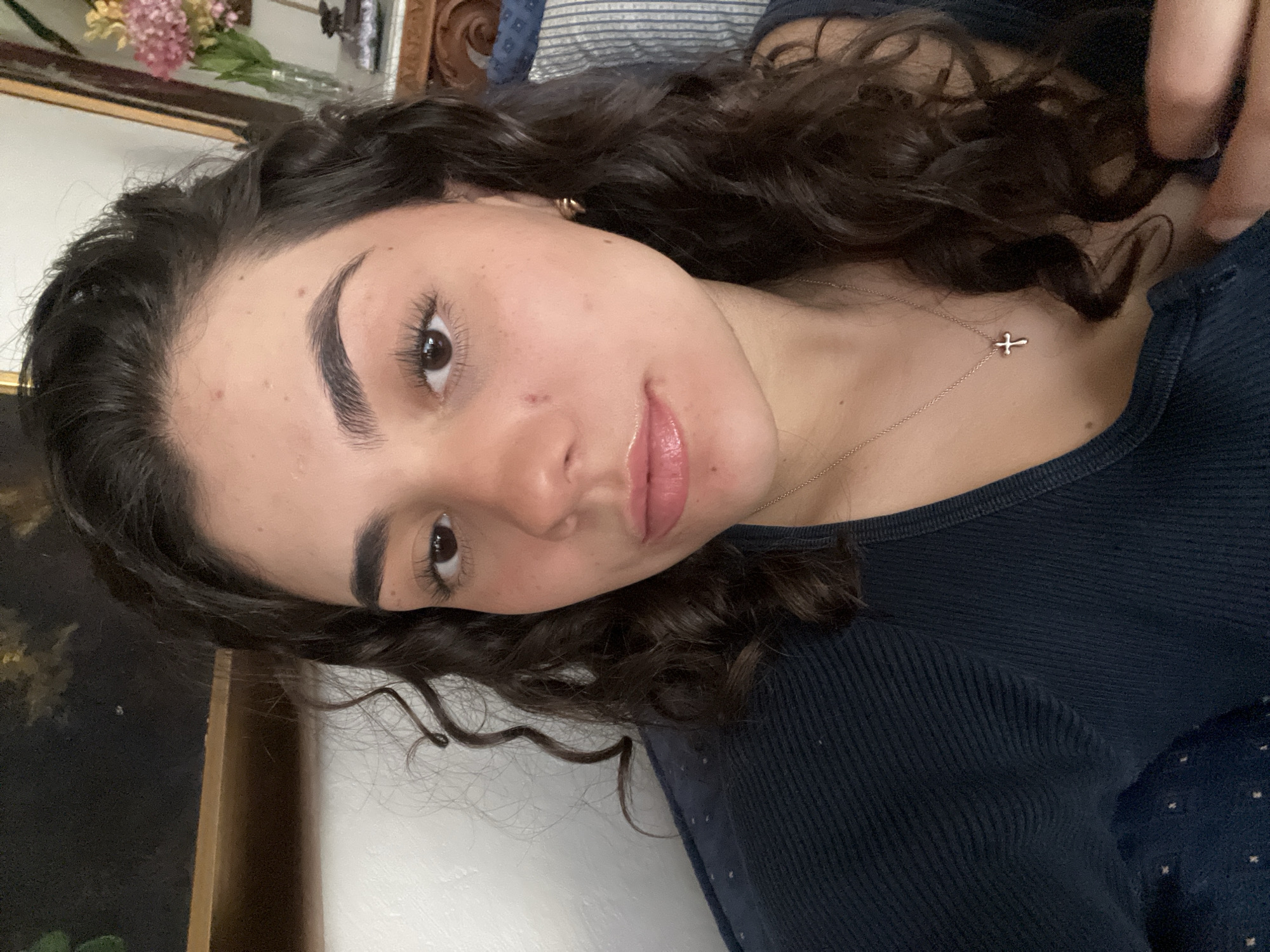 A young woman with curly dark hair and light skin is looking at the camera. She has thick eyebrows and long eyelashes. She is wearing a navy blue ribbed shirt and a delicate gold necklace with a small cross pendant. The background is slightly out of focus, showing a floral arrangement, a wooden frame, and a striped cushion. The location of the scene is unknown.