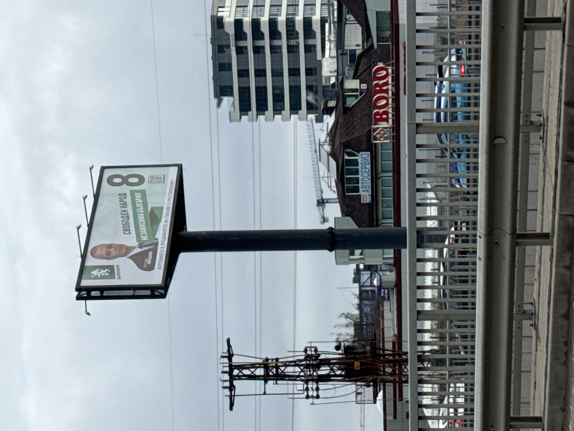 The image shows an outdoor advertising billboard in Sofia, Bulgaria, positioned adjacent to a road or highway. The billboard features a political advertisement for the "Svoboden Narod" (Free People) party. The sign displays the number "8," the party's logo, and a photo of a man, presumably a candidate, and a tagline that warns that the purchase or sale of votes is a crime. The sign is anchored to a dark grey cylindrical pole, and the background includes other structures.
Behind the billboard is a building with a facade of dark-tinted glass and white horizontal detailing, adjacent to a building that houses the “BORO” business as well as a vehicle service center (“АВТОСЕРВИЗ”). Power lines run horizontally across the image. A metal guardrail and an electrical utility structure are in the foreground, suggesting the image was taken from a vehicular overpass or bridge. The sky is overcast, indicating neutral weather conditions.