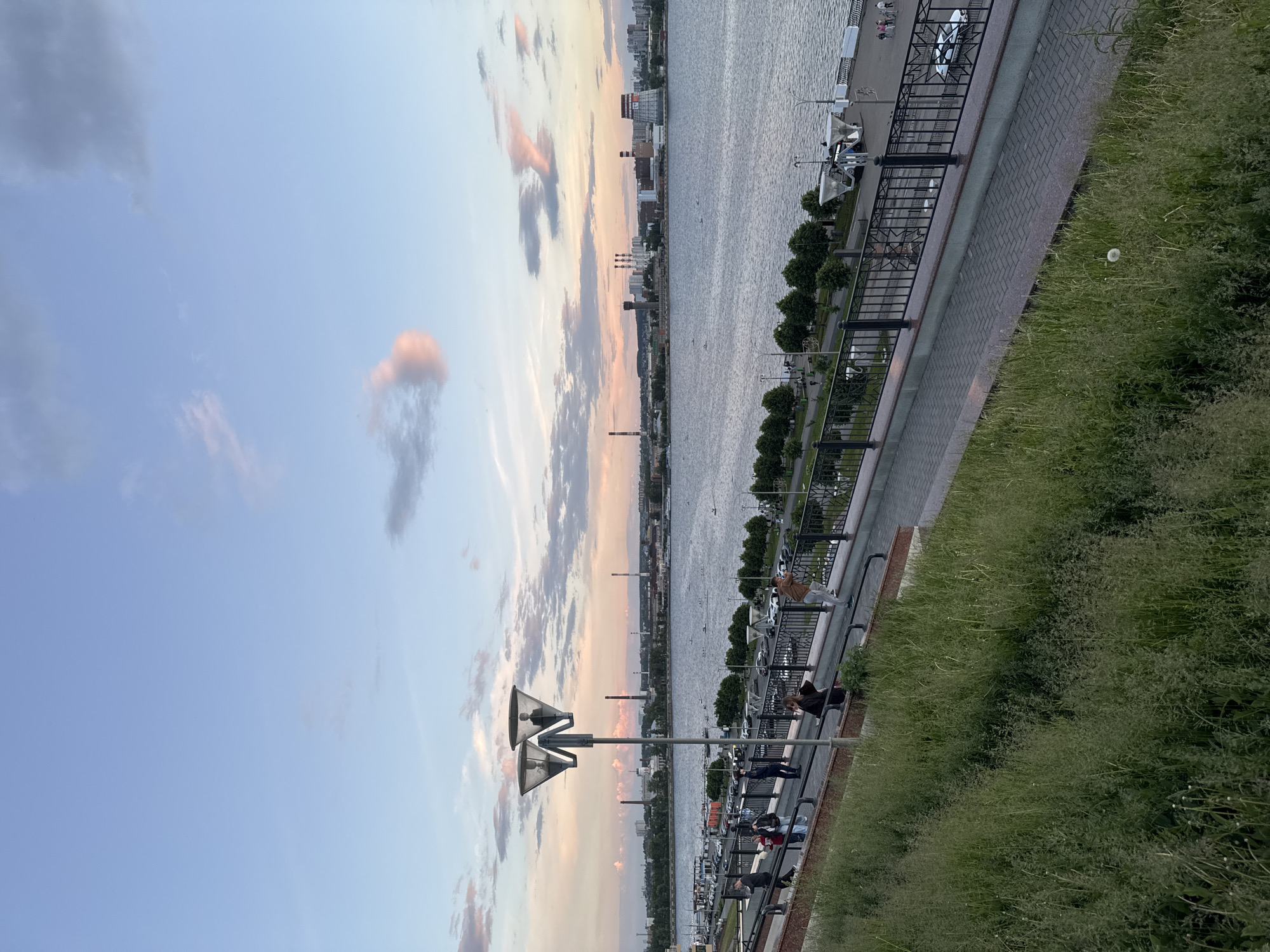 The image captures a serene urban riverfront scene, likely in Izhevsk, Russia, presented in a vertical orientation. The sky, filling the upper portion, transitions from clear blue to scattered clouds touched with soft pink and orange hues, indicative of either sunrise or sunset.
A wide body of water, presumably a river, extends vertically down the left side of the frame, its surface showing gentle ripples. On the distant bank across the river, a city skyline is visible, featuring a mix of buildings and industrial structures, including several prominent chimneys.
Running vertically along the right side of the image is an urban embankment. This comprises a paved walkway, a sturdy dark metal fence with railings, and a sloped bank covered in tall grass. Several casually dressed individuals are present on the walkway, standing or leaning against the fence, seemingly enjoying the peaceful atmosphere. Small trees or bushes are planted along the paved path.
A distinctive street lamp, featuring multiple, possibly reflector-equipped lights, stands prominently in the mid-ground, positioned between the river and the embankment. In the foreground, specifically within the tall grass at the bottom right, a single, fluffy dandelion seed head is visible. The scene conveys a sense of tranquility and leisure in an urban setting during the 'golden hour'. No discernible text is visible in the image.