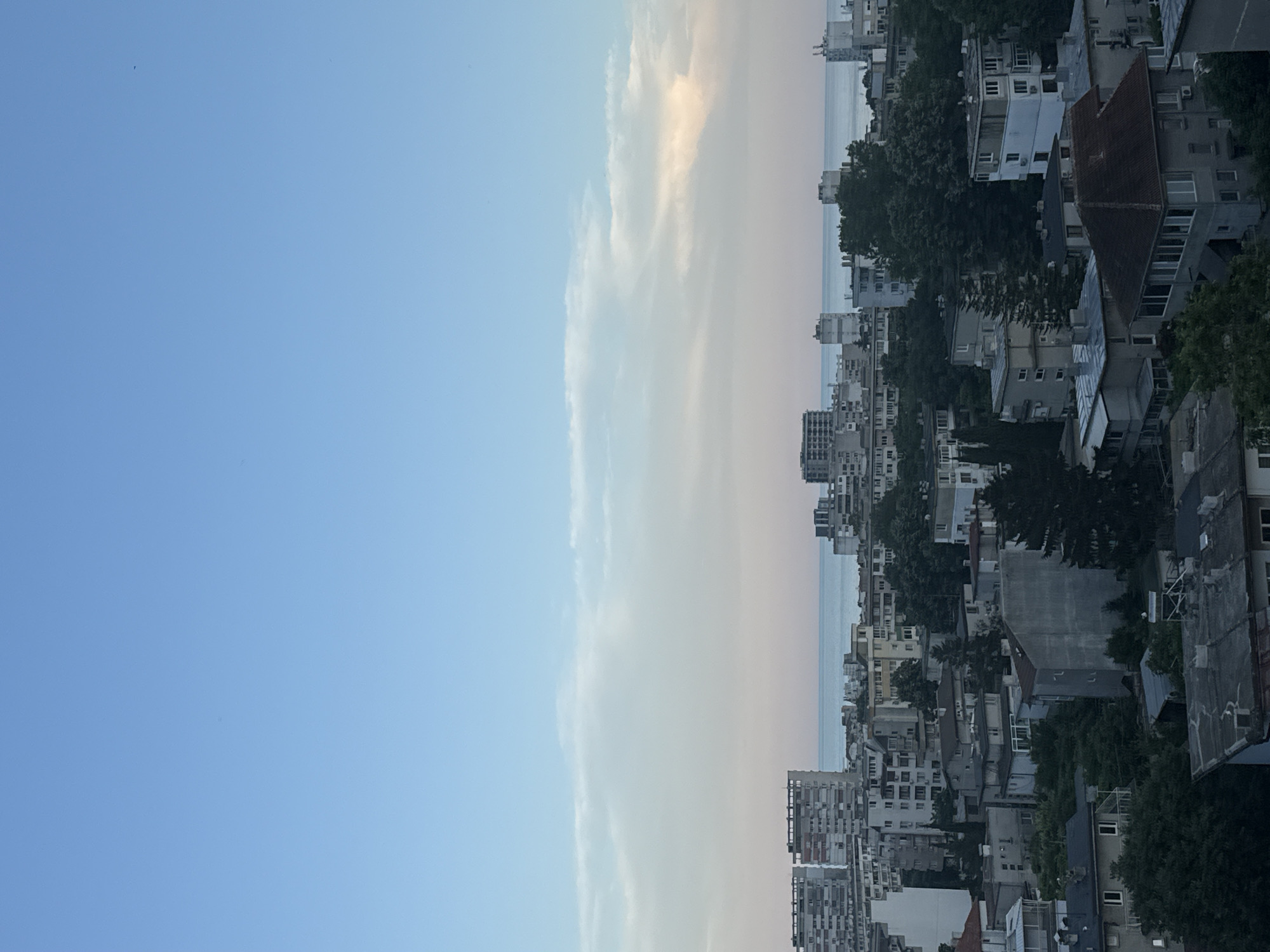 This is an aerial view of a dense urban area with a body of water in the background, likely the Black Sea, as seen from Varna, Bulgaria. The image captures a cityscape dominated by apartment buildings of various sizes and architectural styles, interspersed with trees and greenery. The sky is a soft blue with wispy clouds, transitioning to pink and orange hues near the horizon, suggesting either sunrise or sunset. The buildings are primarily constructed from concrete and show signs of age, with many featuring balconies and visible windows. The overall impression is one of a moderately populated coastal city with a mix of older and more modern residential structures. There are no discernible people or ongoing activities visible in the image.