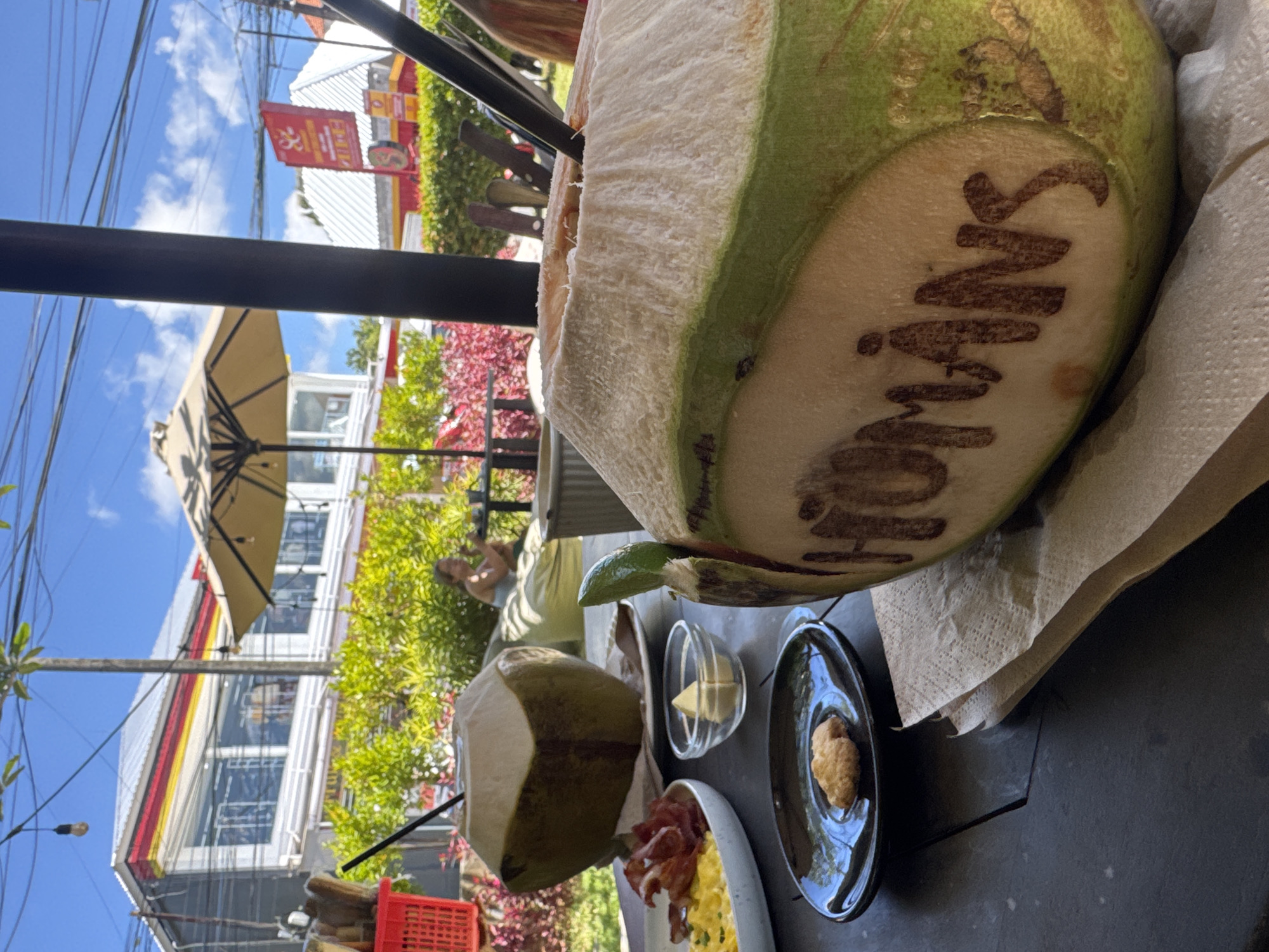 From a high-angle perspective, the image captures a culinary scene at a location in Ungasan, Indonesia. The foreground is dominated by a table setting that includes food and drinks. A partially opened coconut is positioned prominently, with the word "Honians" etched into its surface. Beside it is a plate containing scrambled eggs and bacon, alongside a small bowl containing a pat of butter. A small black plate contains a single cookie.
In the midground, a blurred figure of a person is visible behind the table. The background showcases the outdoor environment of Ungasan, with lush greenery, a building with a red and yellow exterior, and a blue sky dotted with clouds. Parasols provide shade for outdoor seating, and power lines stretch across the sky.