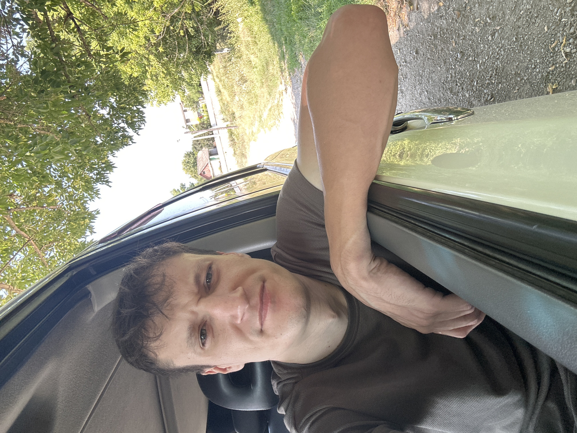 A man is leaning out of a car window, his arm resting on the door frame. He is wearing a brown t-shirt and has short brown hair. The car is parked on a gravel road, with trees and houses in the background. The scene appears to be in a rural area, possibly a village or town. The man's facial expression is neutral, and he is looking directly at the camera. The sunlight suggests it is daytime. The specific location is identified as Makiv, Ukraine, based on the context provided.