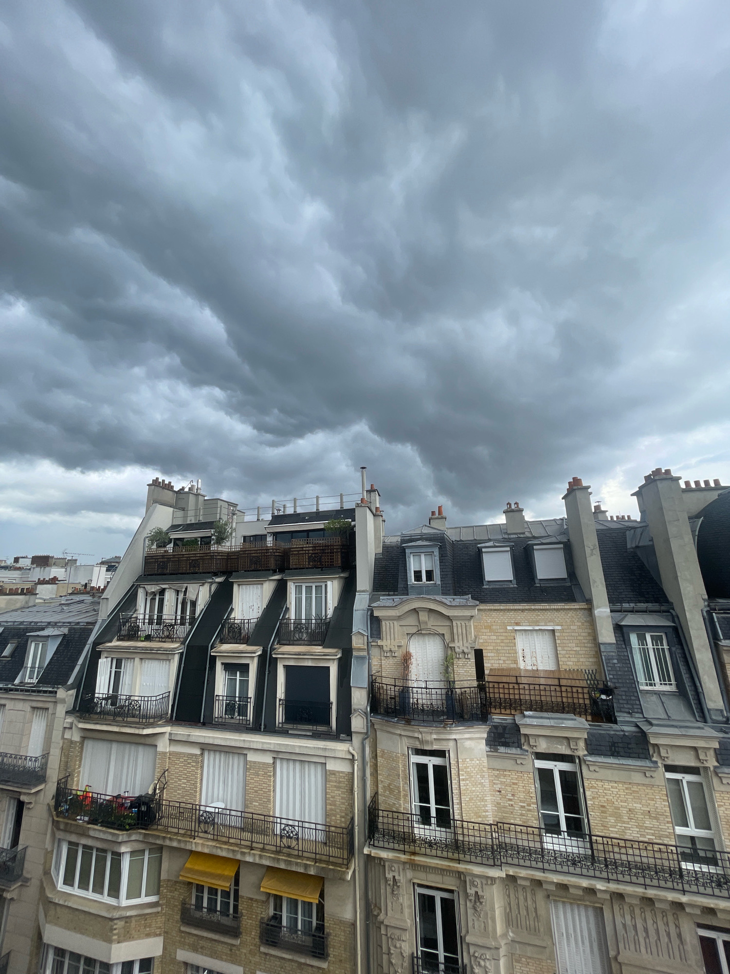 A medium shot captures a dense, overcast sky with dark, textured clouds above a series of Parisian apartment buildings. The buildings feature classic Haussmannian architecture with mansard roofs, dormer windows, ornate balconies with wrought-iron railings, and cream-colored brick facades. Sunlight filters through the clouds, creating a dramatic, somber atmosphere over the cityscape. The buildings display various window types, some with white shutters and others with sheer curtains. Small balconies are visible, some adorned with plants and outdoor furniture. Chimneys punctuate the rooftops, and satellite dishes are present on some of the upper levels. The scene appears to be from an elevated vantage point, possibly a higher floor of a building, looking down upon the adjacent structures and towards the ominous sky.