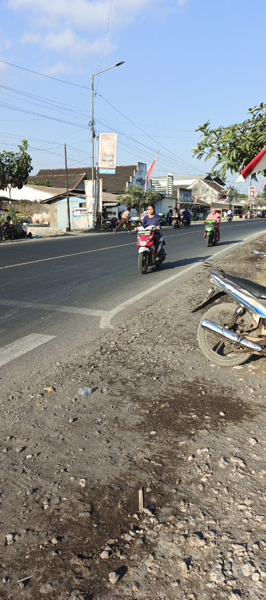 A sunny day scene on a roadside in Kasiyan, Indonesia. The image focuses on a paved road with a rough shoulder made of dirt and gravel. A woman in a blue shirt rides a red and white scooter in the middle of the road. To her right, another woman in a pink headscarf rides a green scooter. In the background, there are buildings, streetlights, and more scooters. The sky is clear and blue.